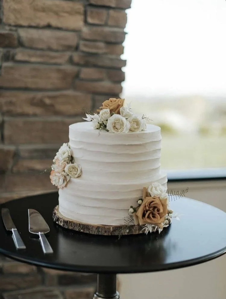 White three-tier wedding cake decorated with beige and white roses, placed on a black cake stand with cake cutting utensils beside it, in front of a brick wall and window.