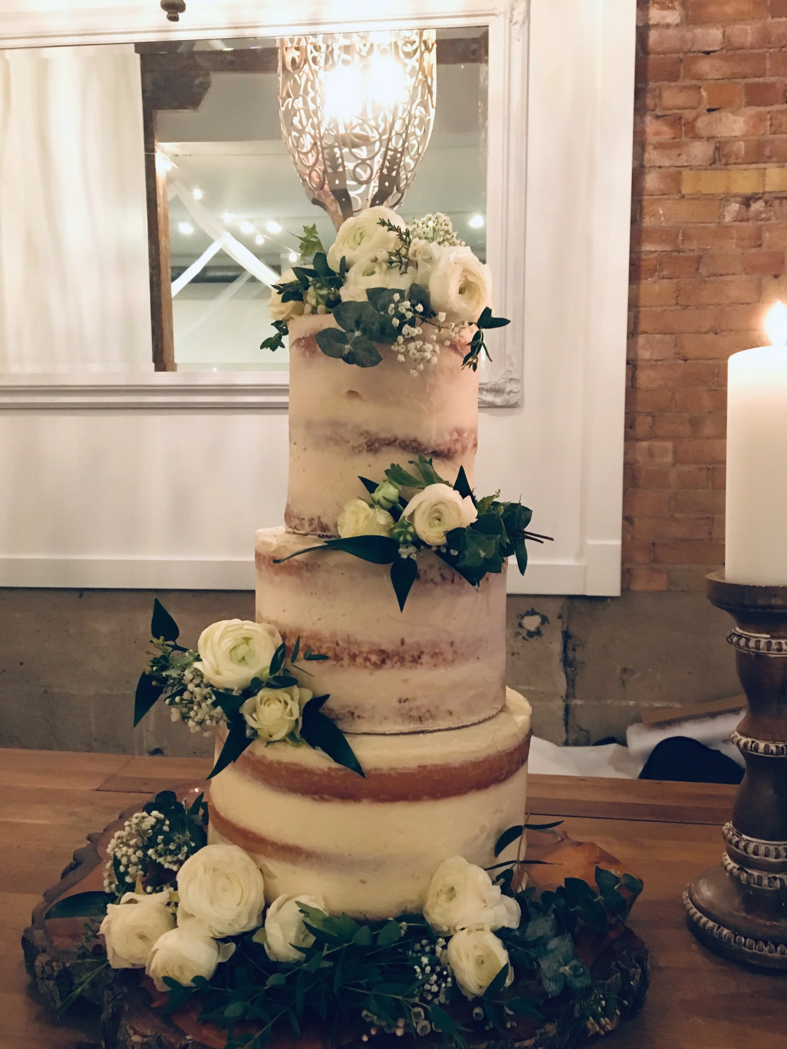 A multi-tiered wedding cake decorated with white roses, baby's breath, and green foliage, placed on a table with additional flowers and greenery.