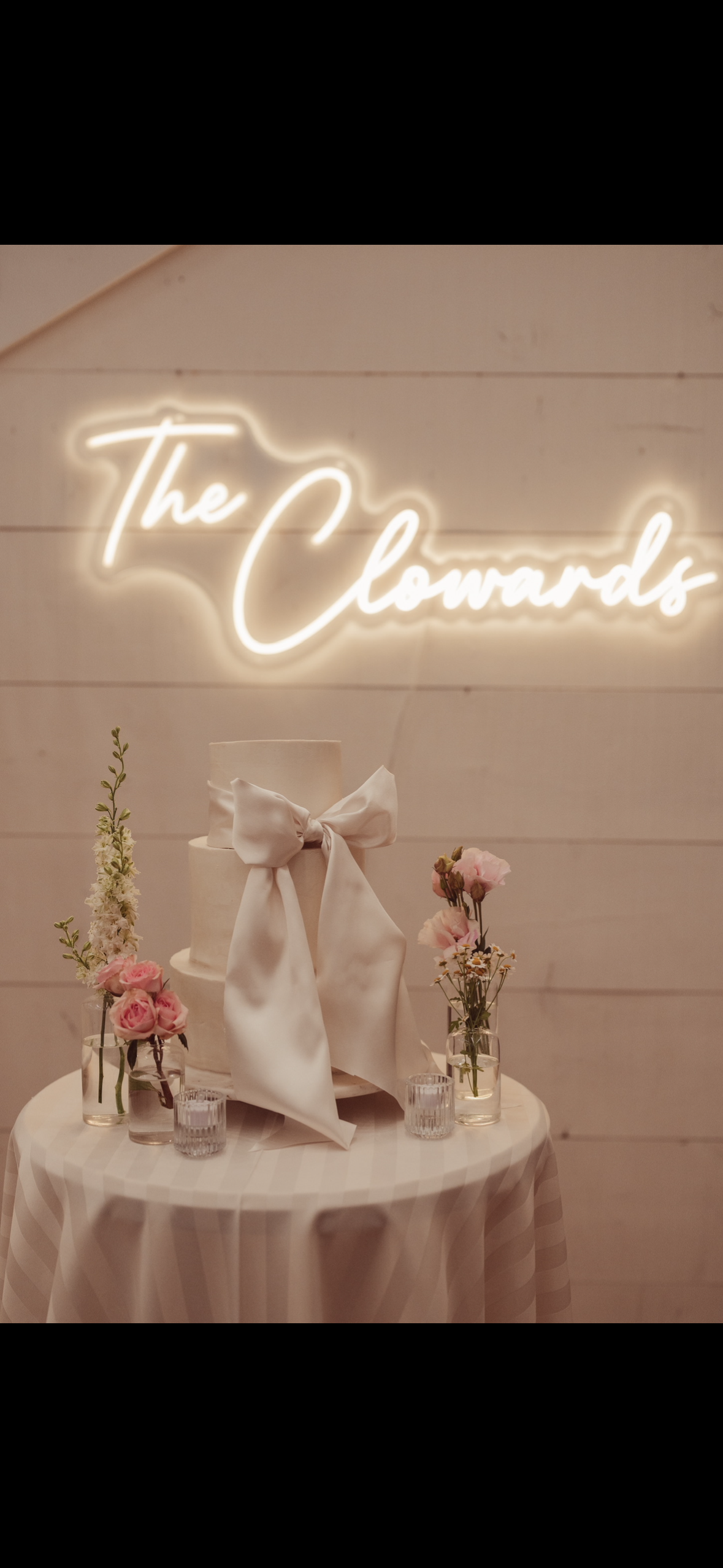 A decorated table with pink flowers in vases, a white cake with a large bow, and a neon sign that says 'The Cheers' in the background.