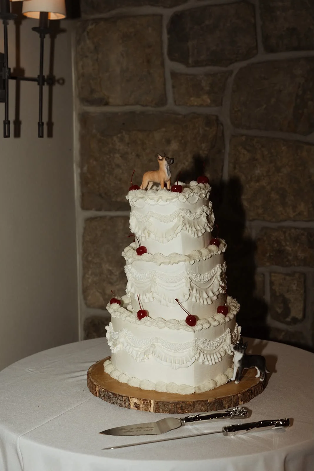 A three-tiered white wedding cake with ruffled icing and cherries on top, placed on a round wooden cake stand on a table with a white tablecloth. Two small dog figurines, one on top of the cake and one at the base, flank the cake. A cake knife and server are on the table in front of the cake. The background features a stone wall and wall-mounted lamps.