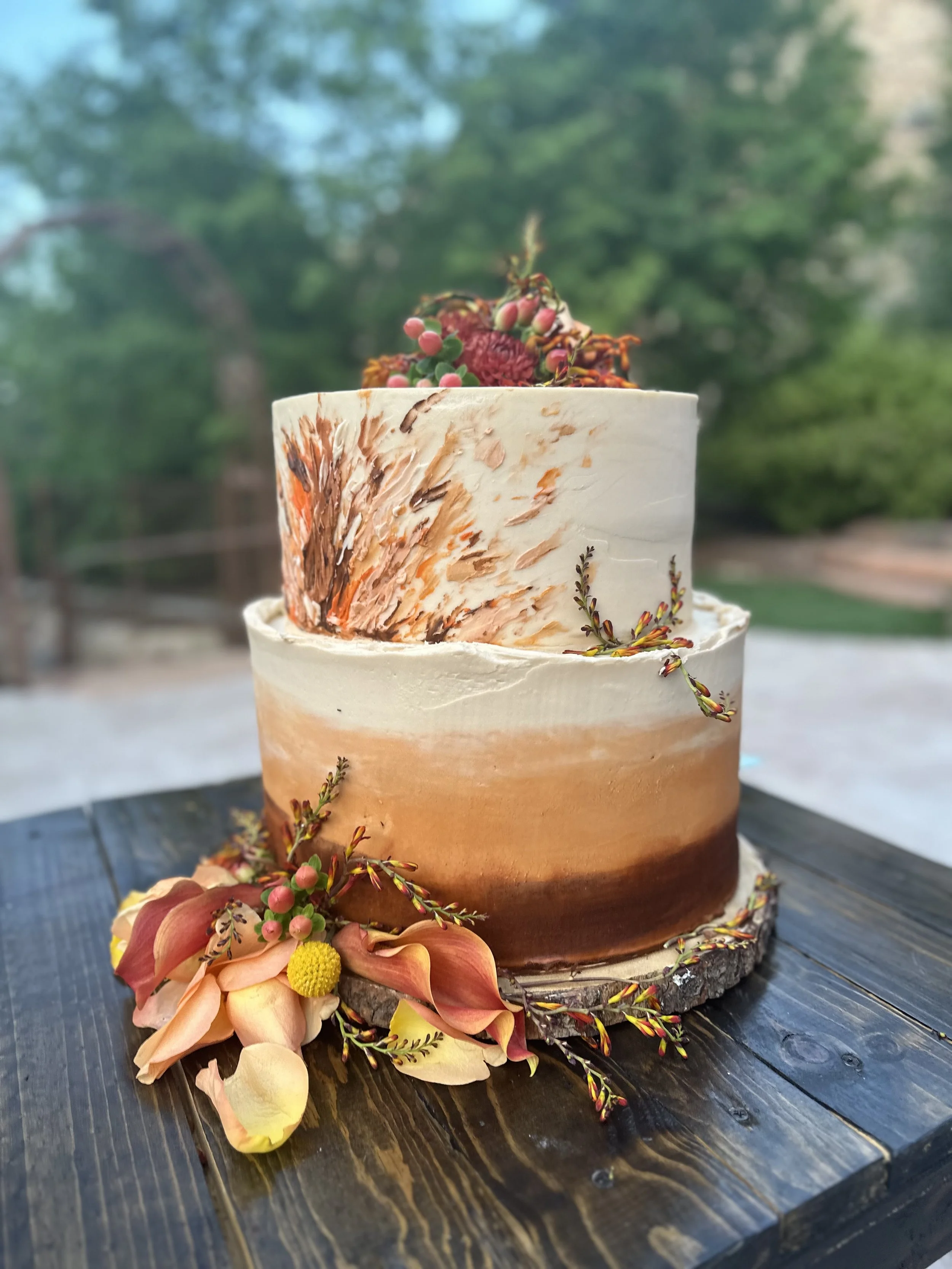 A tiered cake with a wood grain pattern and natural decorations, set on a wooden surface outdoors.