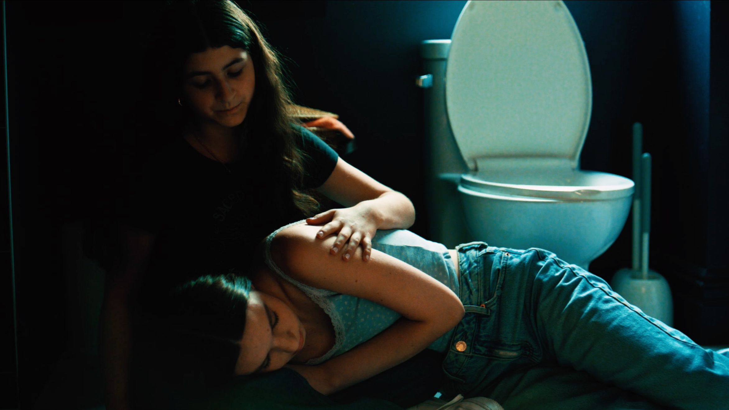 A young woman giving a lap dance to a man lying on the floor in a dimly lit bathroom with a toilet in the background.