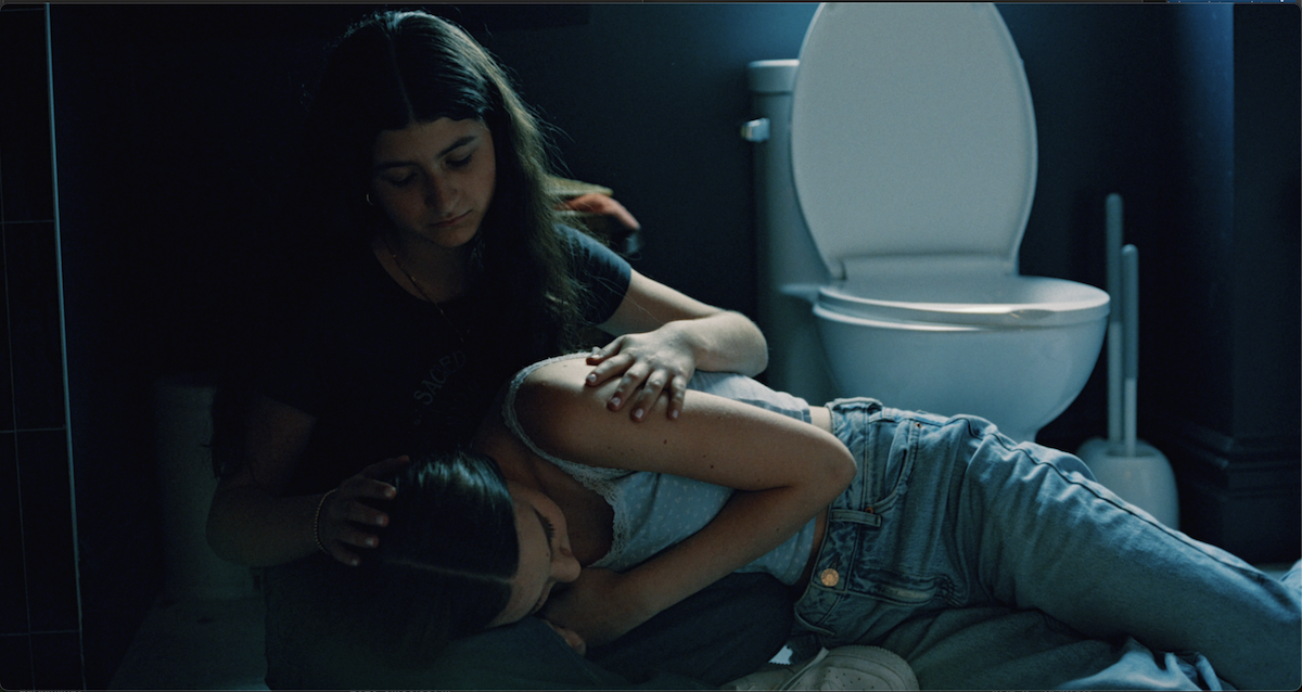Zara and MIla in a bathroom, one sitting on the floor and the other leaning over her.