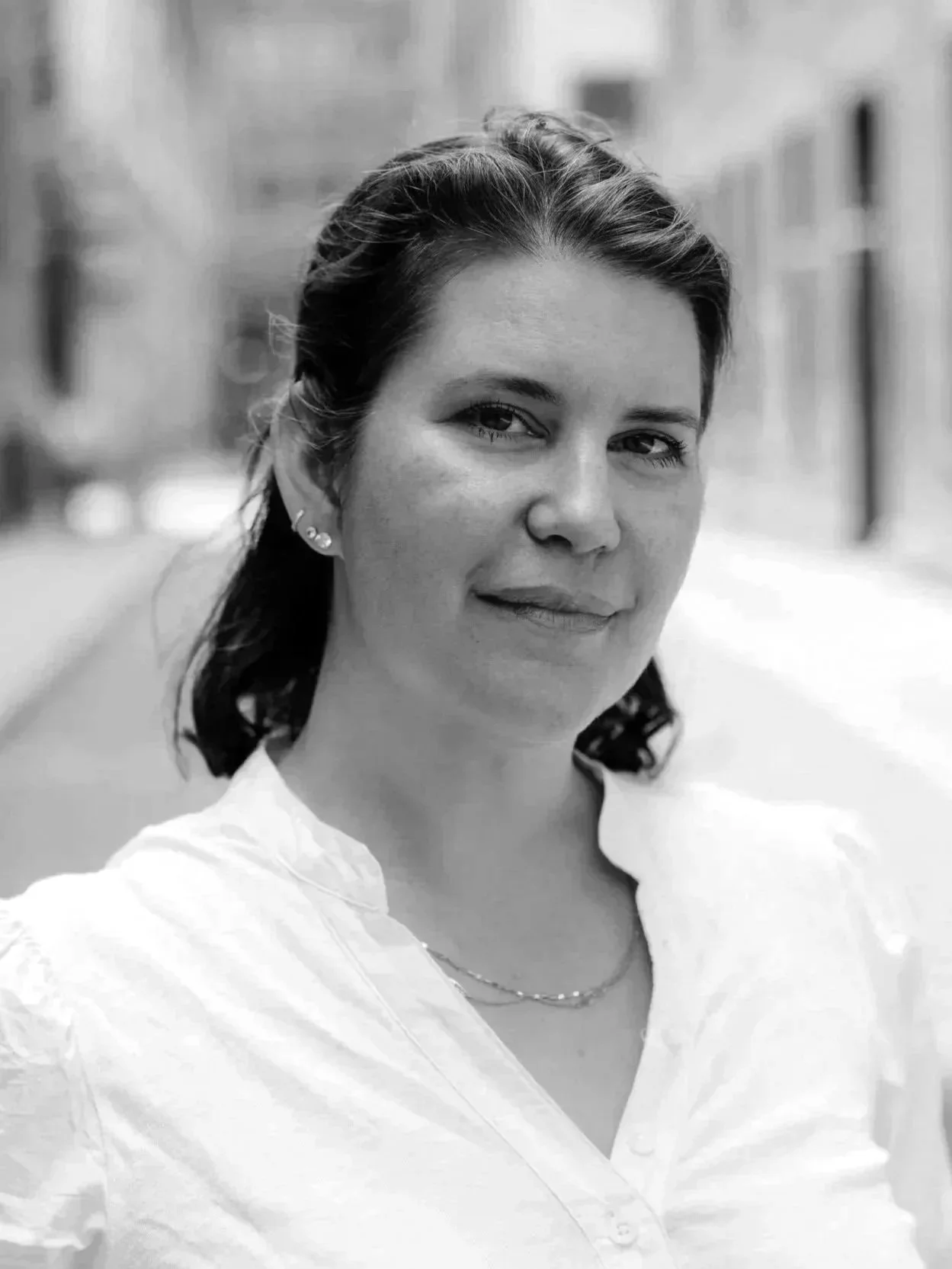 A black and white portrait of a woman with dark hair, wearing earrings and a white top, standing outdoors in an urban setting.