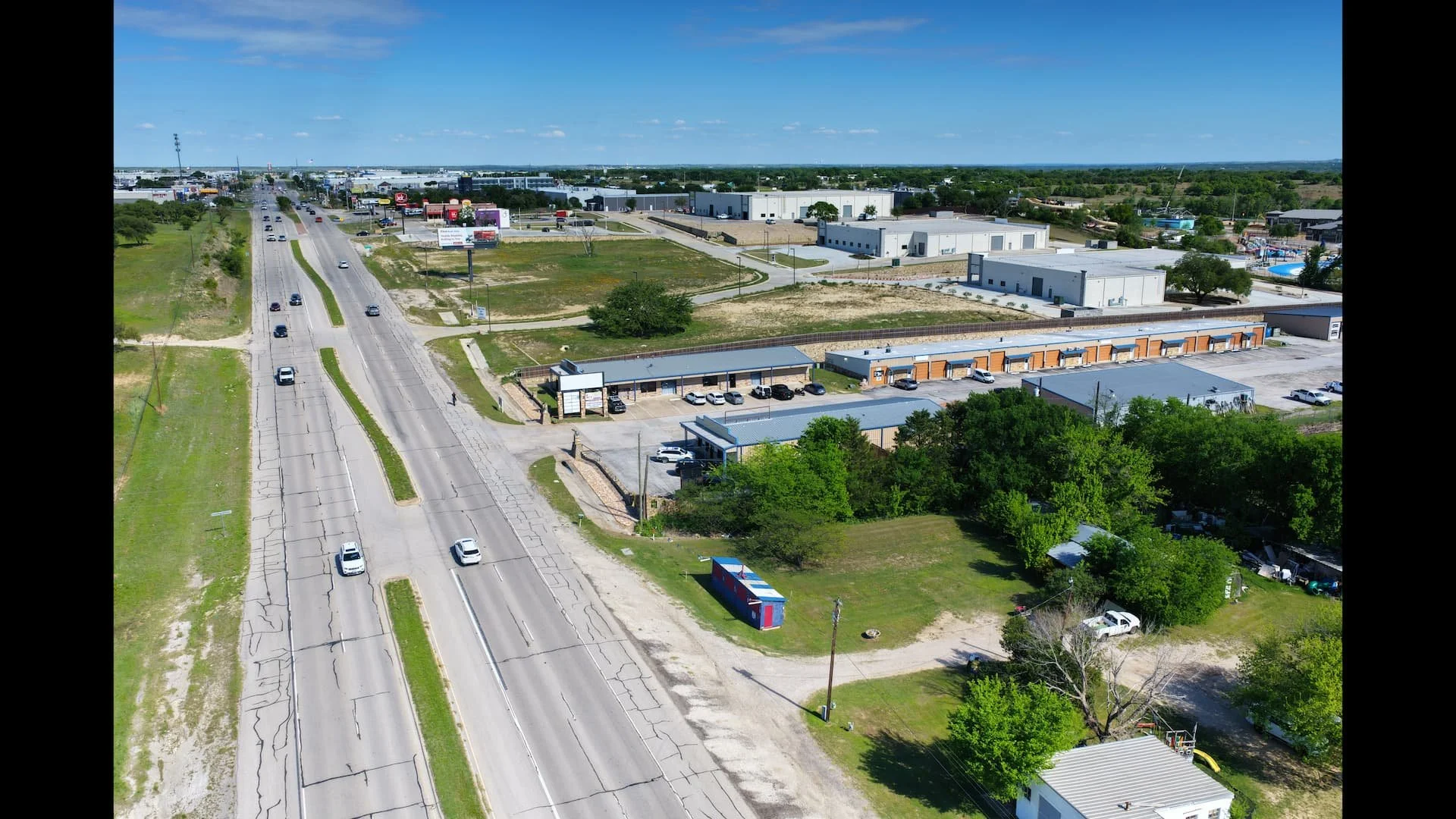 Aerial view of a commercial area with several warehouses and retail stores, adjacent to a busy street with multiple cars, on a sunny day with clear blue skies.