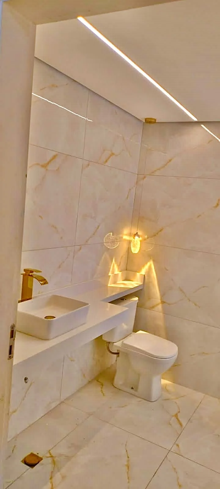 Banheiro com paredes e piso de mármore com detalhes dourados, pia quadrada sobre bancada branca, torneira dourada, vaso sanitário branco e iluminação moderna com lâmpada exposta e iluminação embutida no teto.