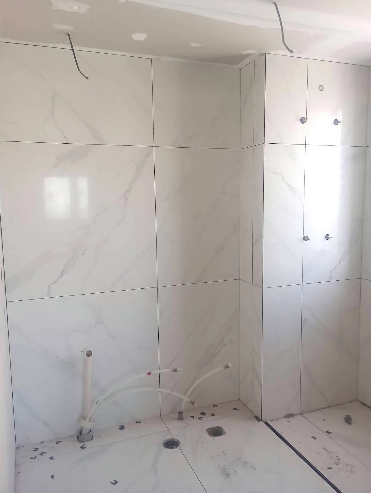Sala de banheiro em construção com paredes revestidas de azulejos brancos com veios cinza, sem equipamentos instalados, com tubos aparentes na parede e buracos no piso para futuras instalações hidráulicas.
