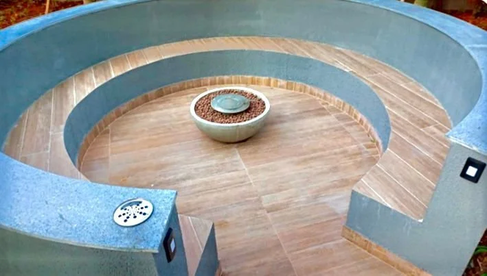 Piscina de forma circular com duas áreas de nível diferente, revestida com azulejos azuis e piso de madeira, contendo uma cuba decorativa no centro cheia de pedras decorativas.