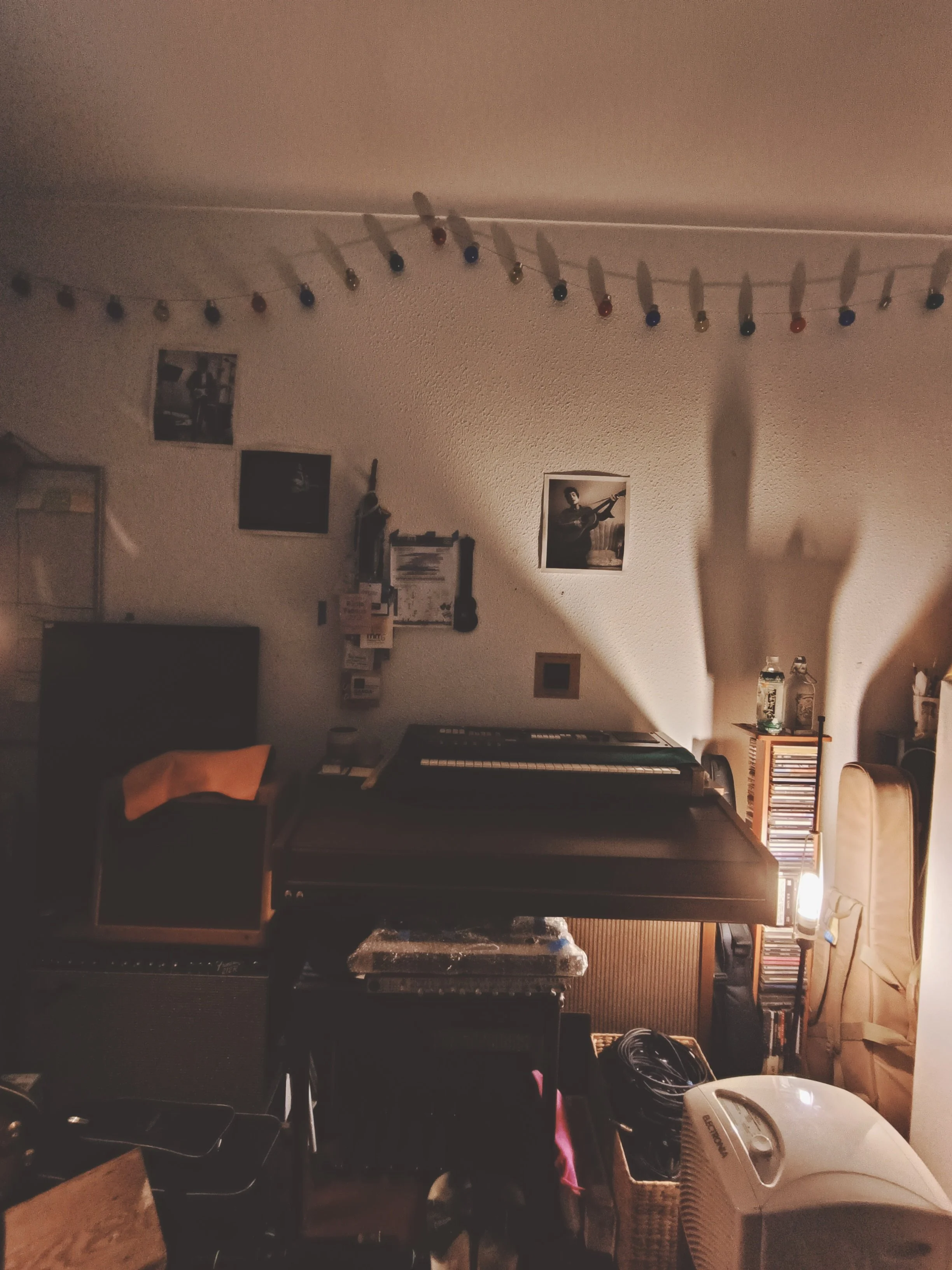 Dimly lit indoor room with a wall decorated with framed photos and posters, a keyboard, various furniture, a fan, and household items.