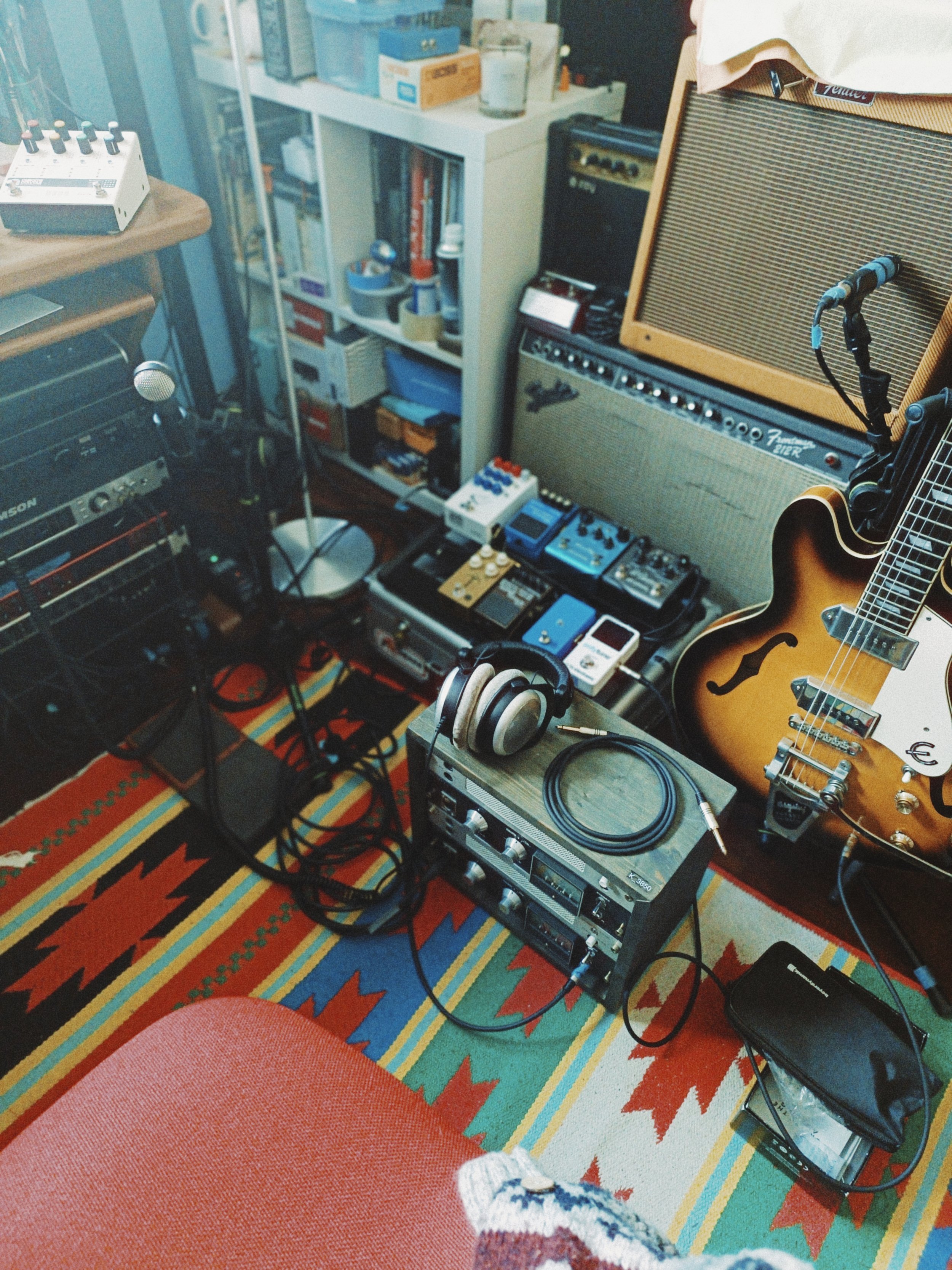 A cluttered music studio space with a guitar, guitar amplifier, effects pedals, overhead microphone, headphones, and various audio equipment on a colorful patterned rug. There is a bookshelf with books, boxes, and stationery in the background.