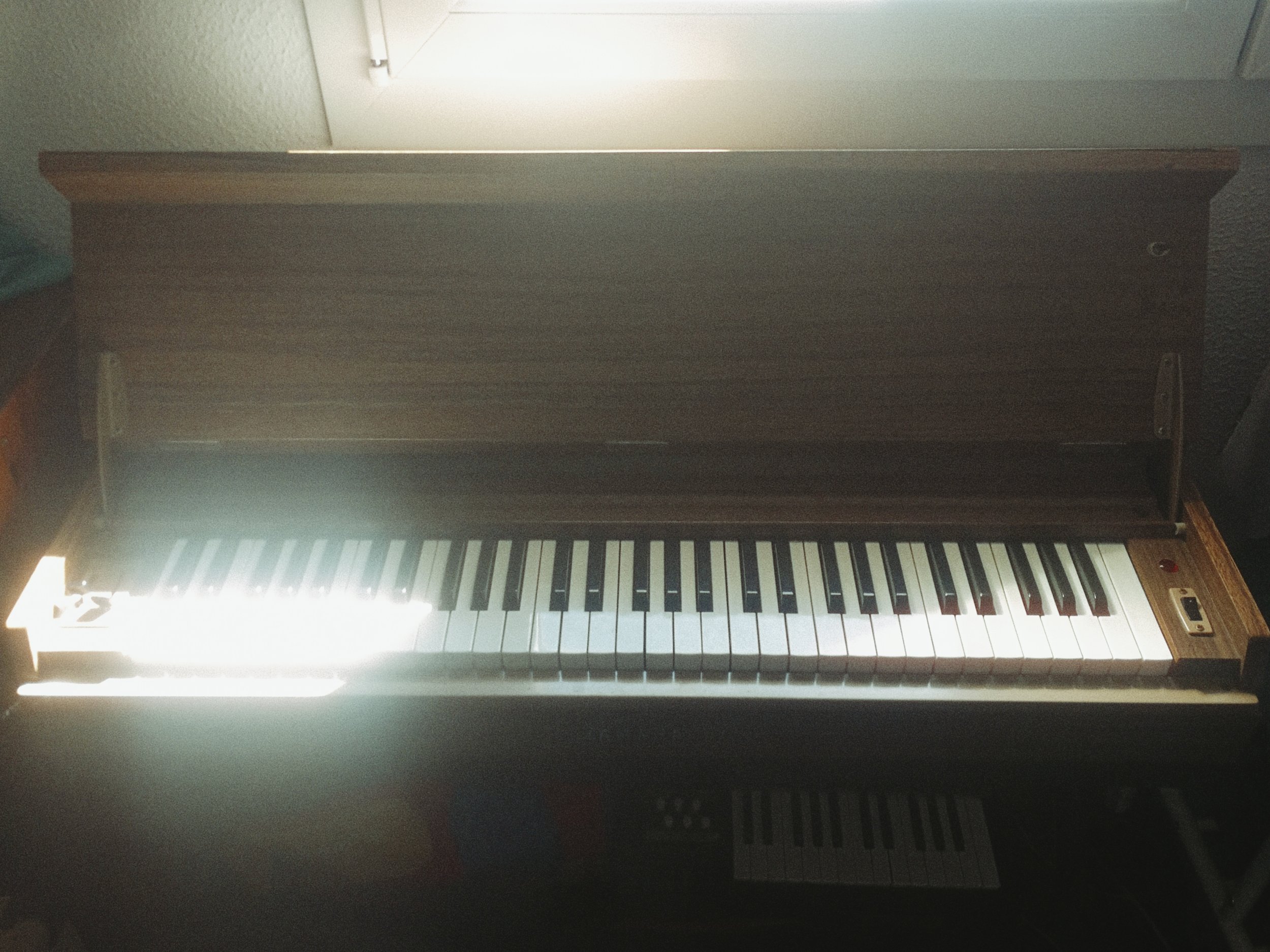 The image shows the keys of a piano with a music stand behind it and a light above illuminating the area.