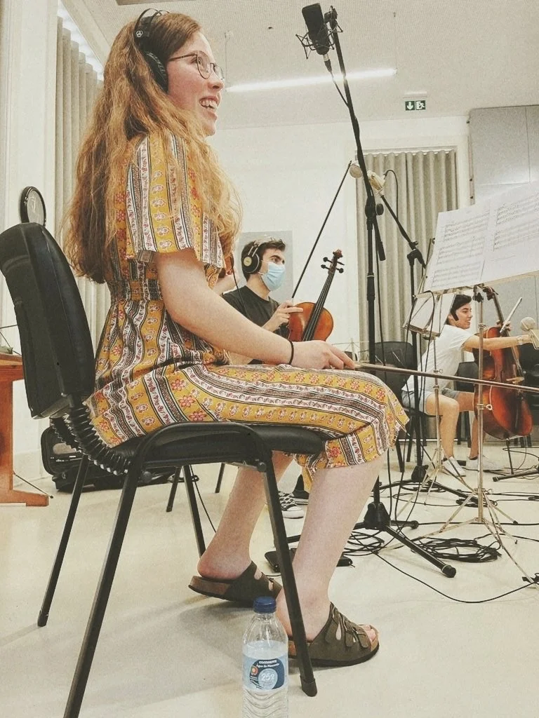 Young woman with long red hair wearing glasses and a patterned dress, sitting on a black chair with a water bottle on the floor, playing a musical instrument in a recording studio. Behind her, a young man wearing a face mask and headphones is playing