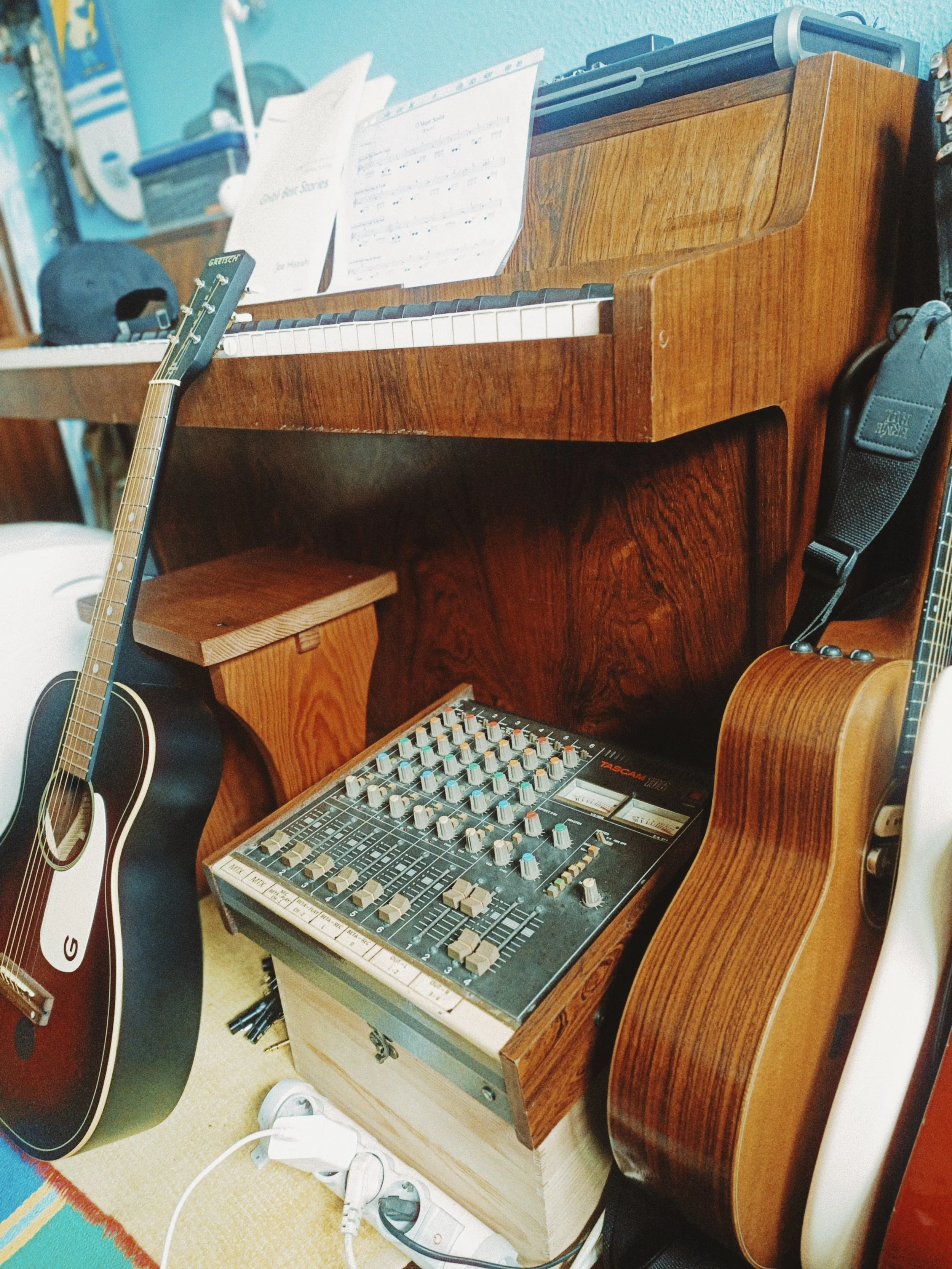 A room with a wooden desk holding sheet music, a black guitar, a wooden stool, an audio mixing console, and a wooden guitar leaning against the desk, with various cables and electronic devices on the floor.