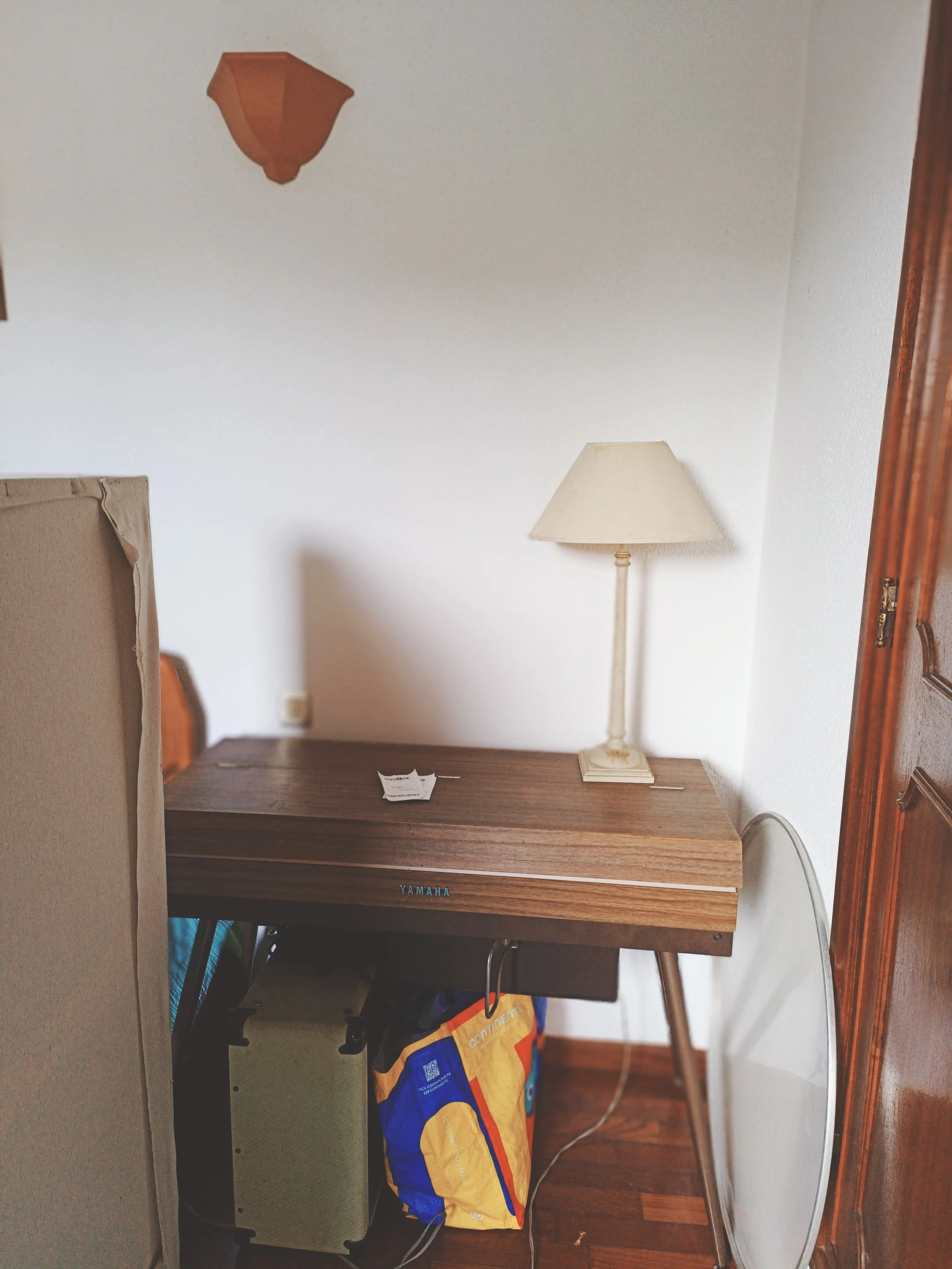 A vintage Yamaha electric piano in a small room, with a table lamp on the right, a cardboard box on the left, and a backpack under the table, against a white wall with a power outlet.