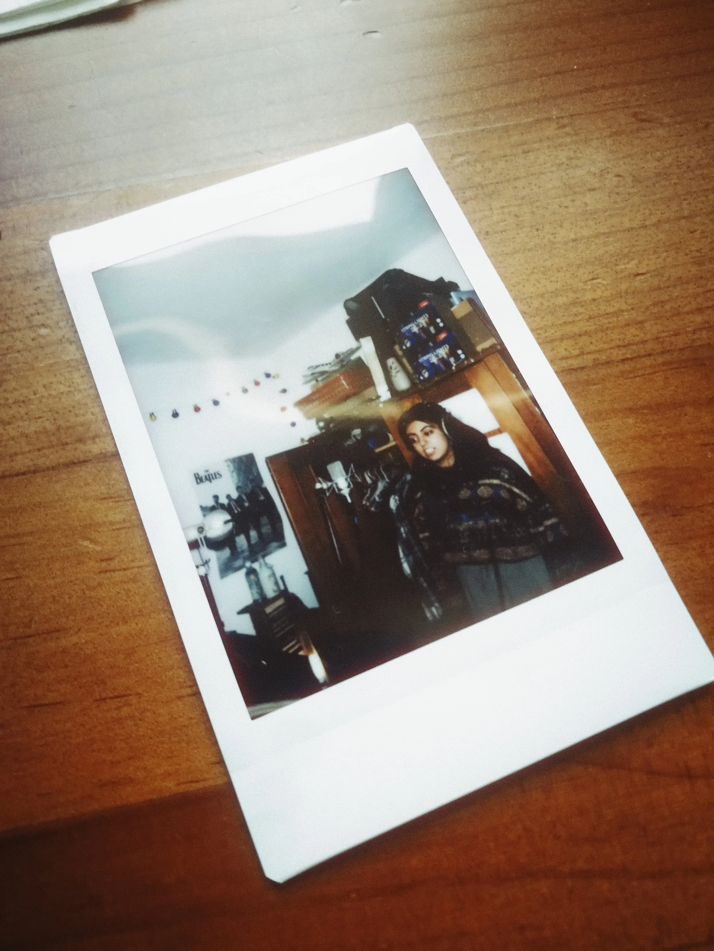 Photograph of a printed photo on a wooden surface showing a woman with dark hair and headphones, standing in a room with shelves, boxes, and wall decorations.