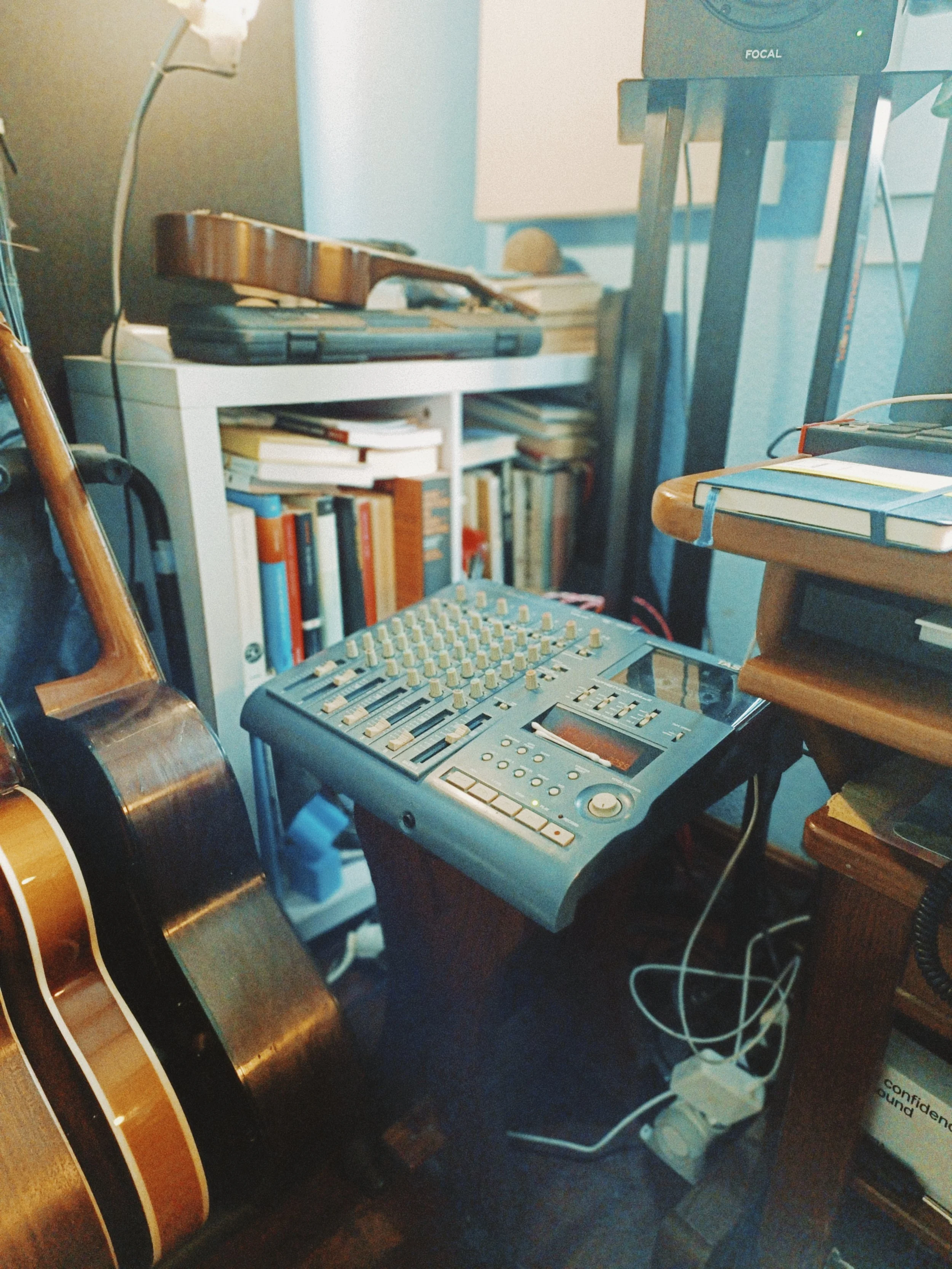 A room with a guitar, a keyboard, bookshelves with books, and some electronic equipment on a desk. There are wires and a power strip on the floor.