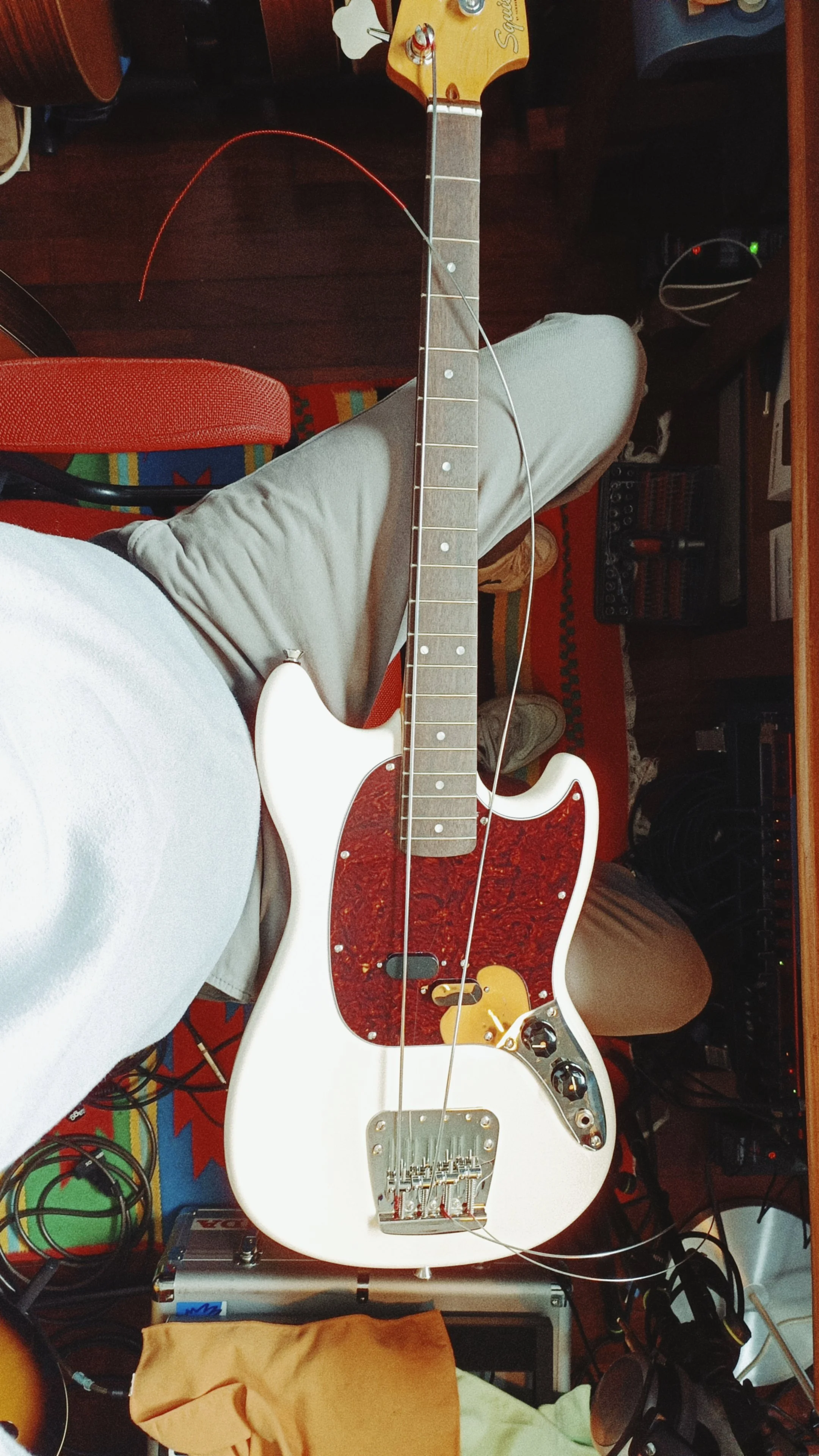 A white electric guitar with a tortoiseshell pickguard and yellow knob, resting on a wooden surface in a room with various electronic equipment and a colorful rug.