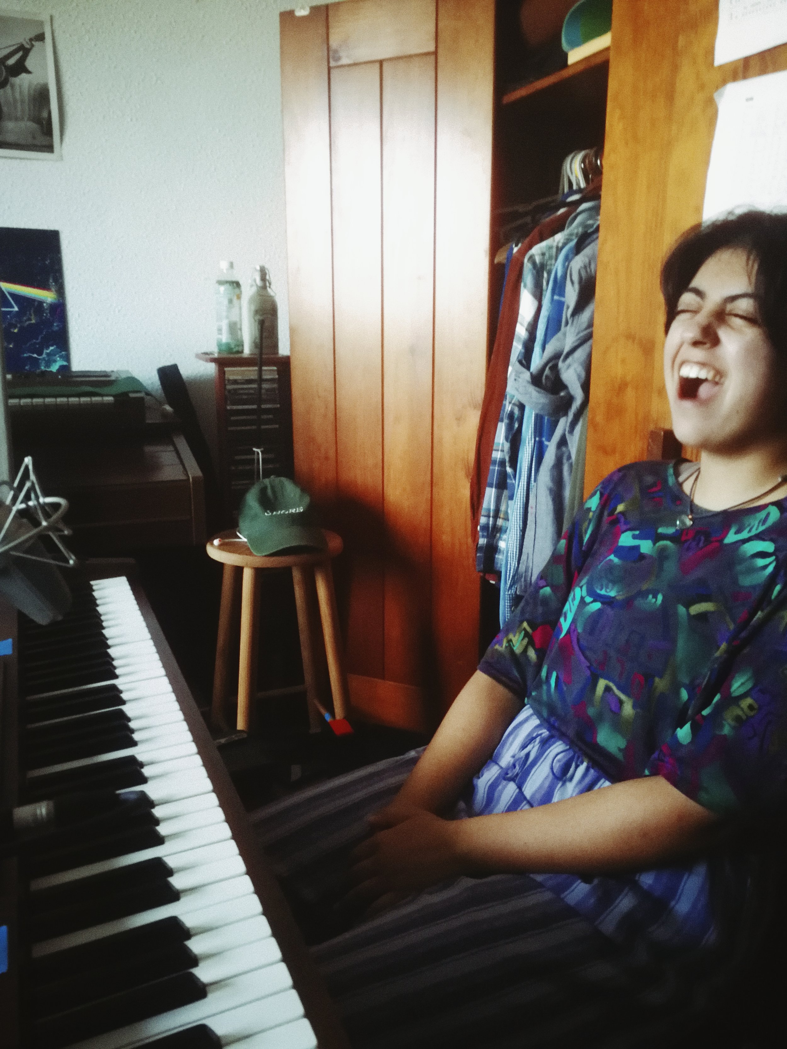 A person sitting at a digital piano keyboard, laughing with eyes closed, wearing a colorful shirt and striped pants, in a room with a wooden wardrobe, clothes hanging inside, a small table with a green cap on it, and bottles on top of a small dresser