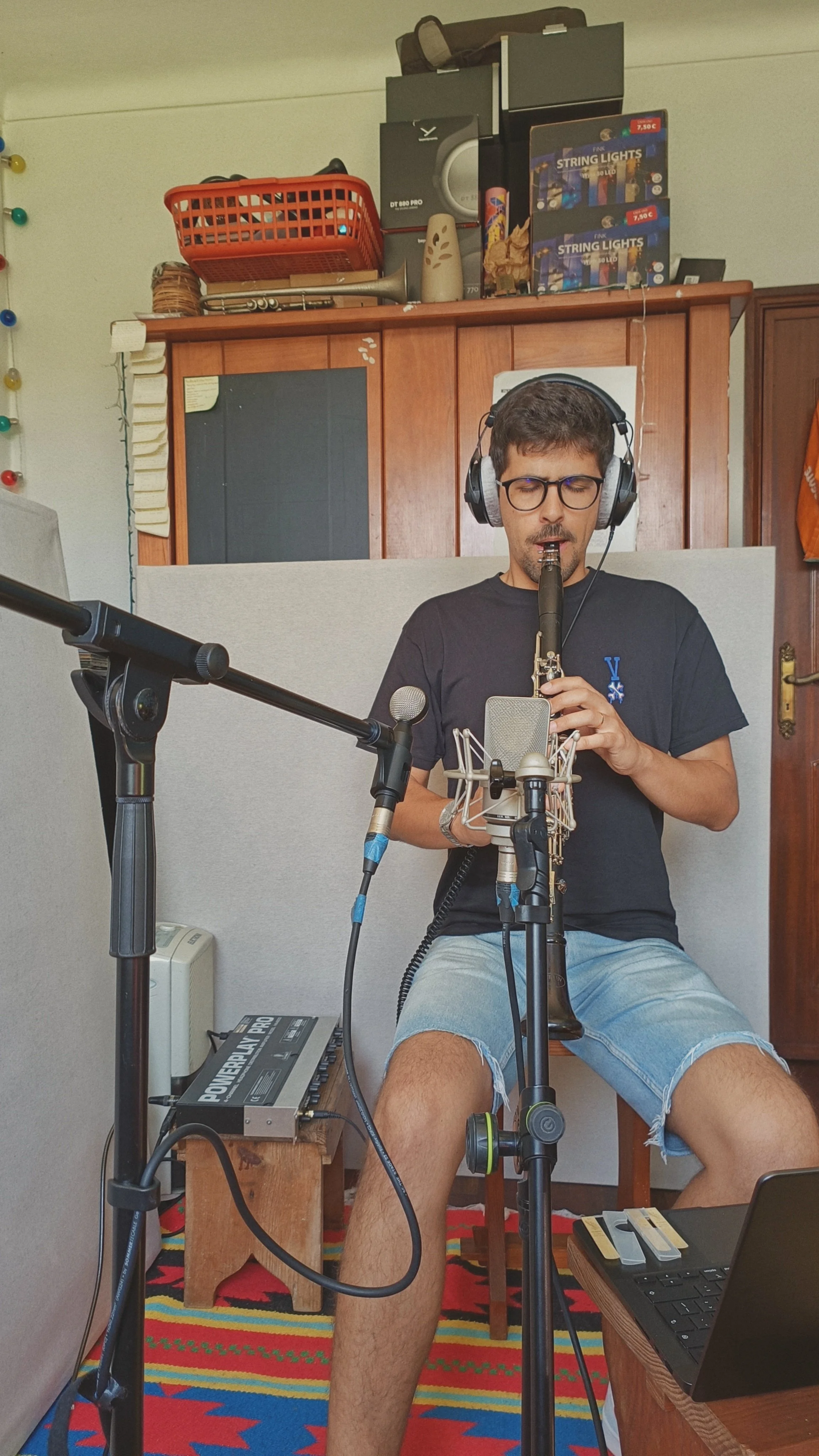 A man wearing glasses and headphones sitting in a room, playing a wind instrument, surrounded by music equipment and furniture.