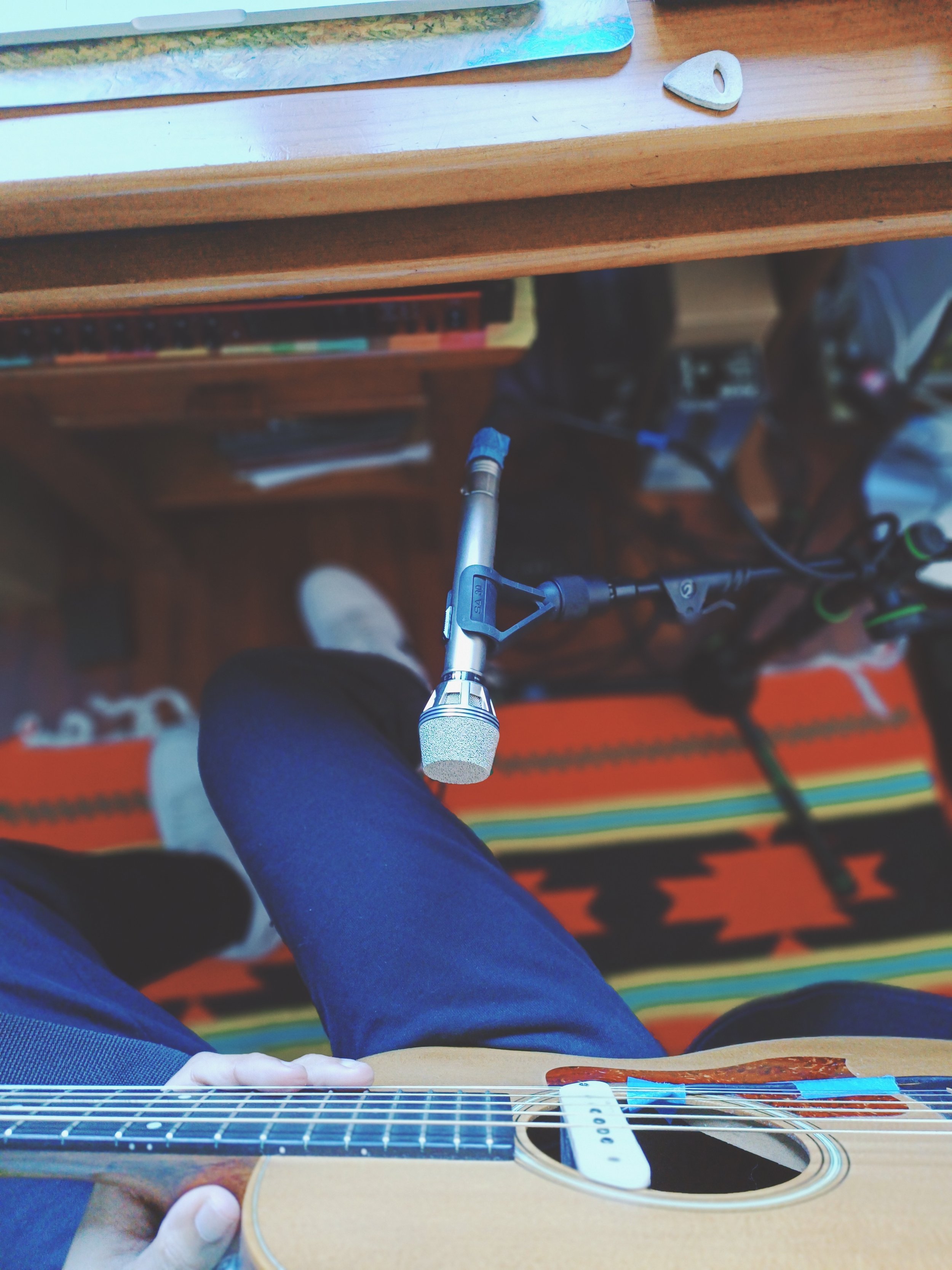 Person playing an acoustic guitar, viewed from above, with a microphone in front of them, and a shelf with books and other objects in the background.