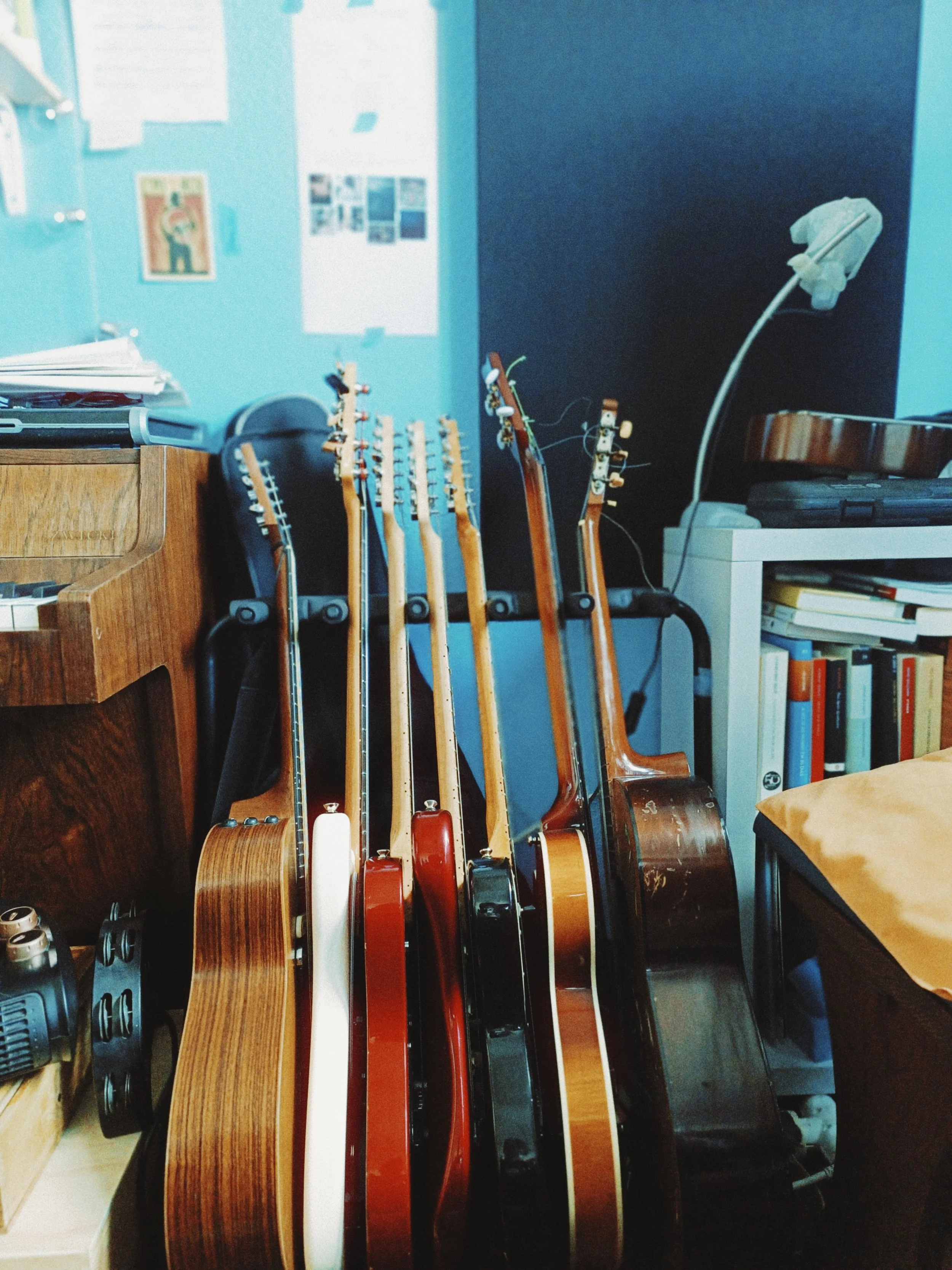 A collection of guitars with different colors and wood finishes placed vertically against a blue wall in a room with posters and documents on the wall, a small white bookshelf with books, and various objects including a power drill and a lamp.