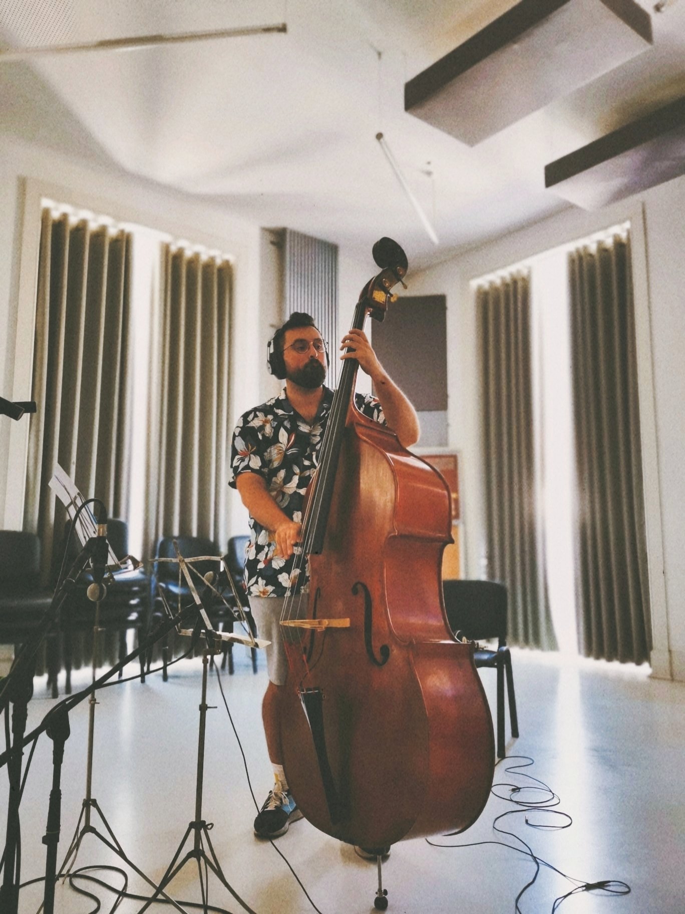 A man wearing glasses and a Hawaiian shirt playing a double bass in a recording studio, with multiple microphones set up around him and soundproof curtains on the walls.