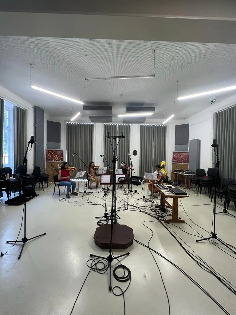 A small group of musicians practicing in a room with microphones, music stands, and chairs, with some musical instruments on stands and cables on the floor.