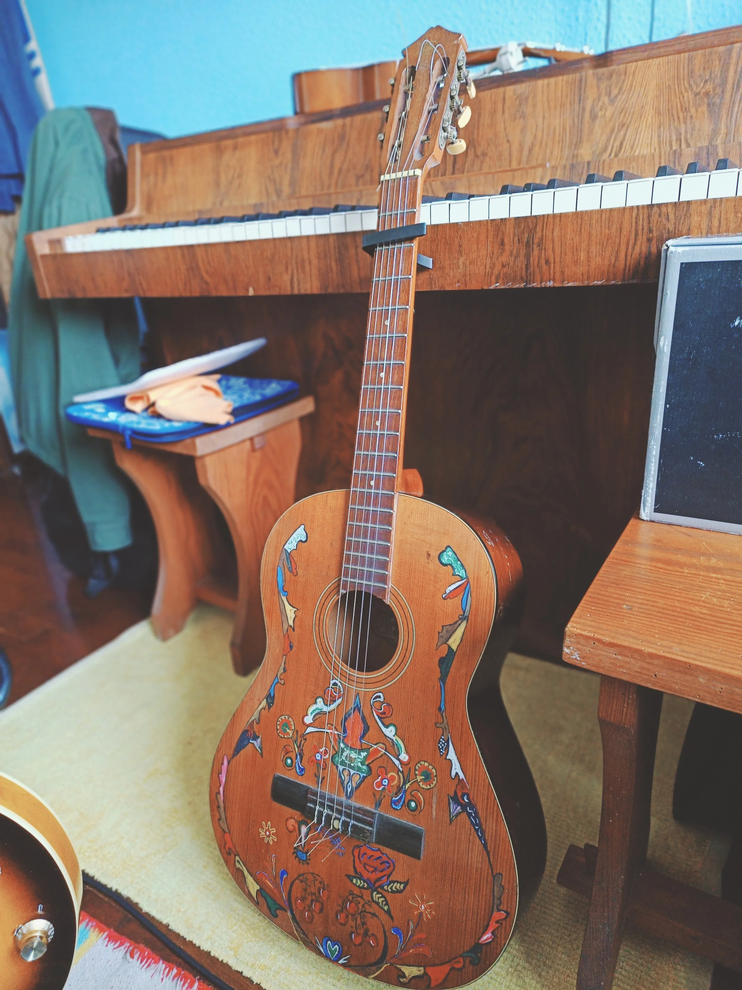 A wooden acoustic guitar with colorful painted designs on its body, leaning against a wooden table in a room with a blue wall.