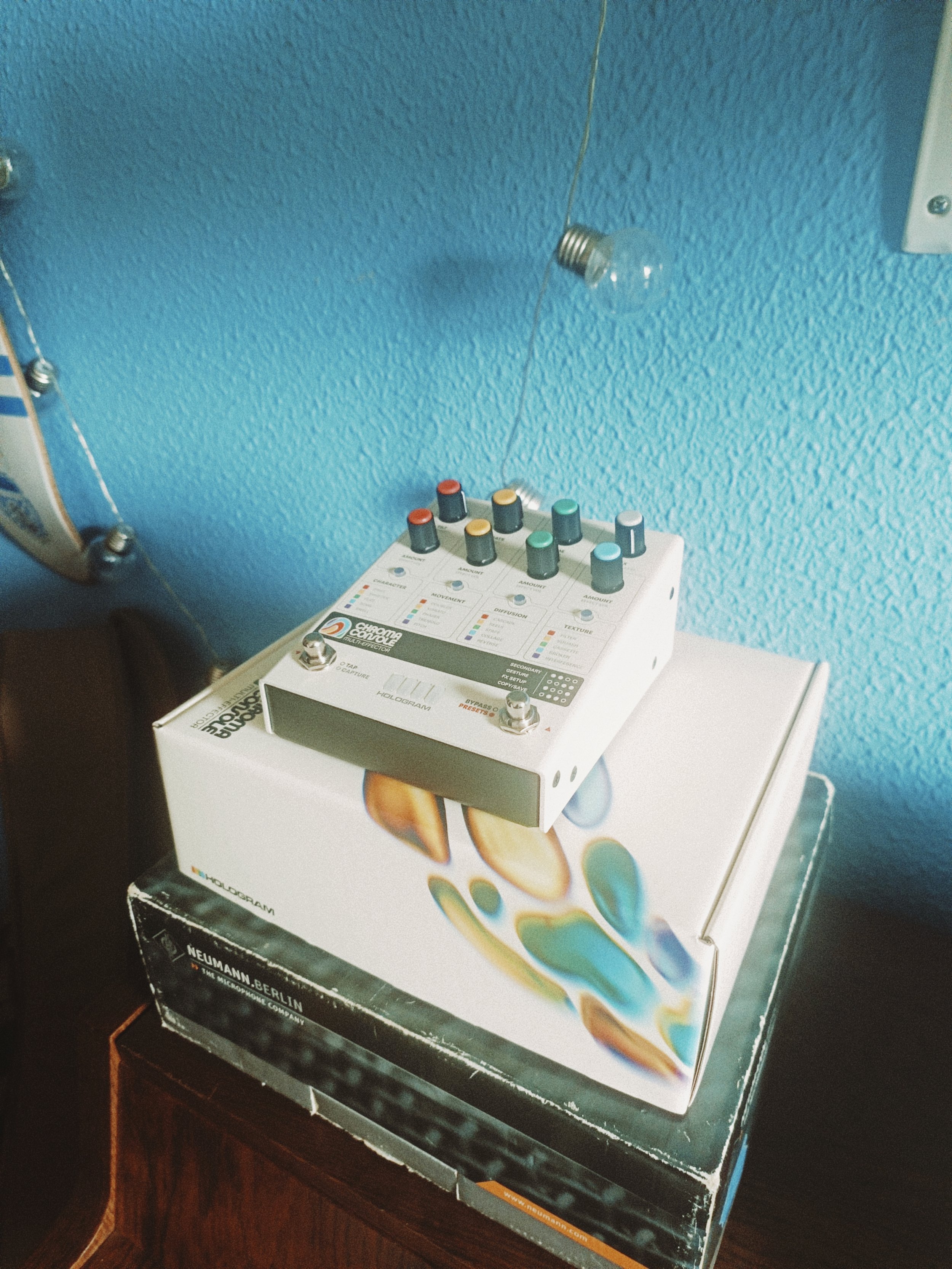 A musical effects pedal on top of its box, which is placed on a wooden table. The pedal has multiple knobs and switches for controlling various effects.