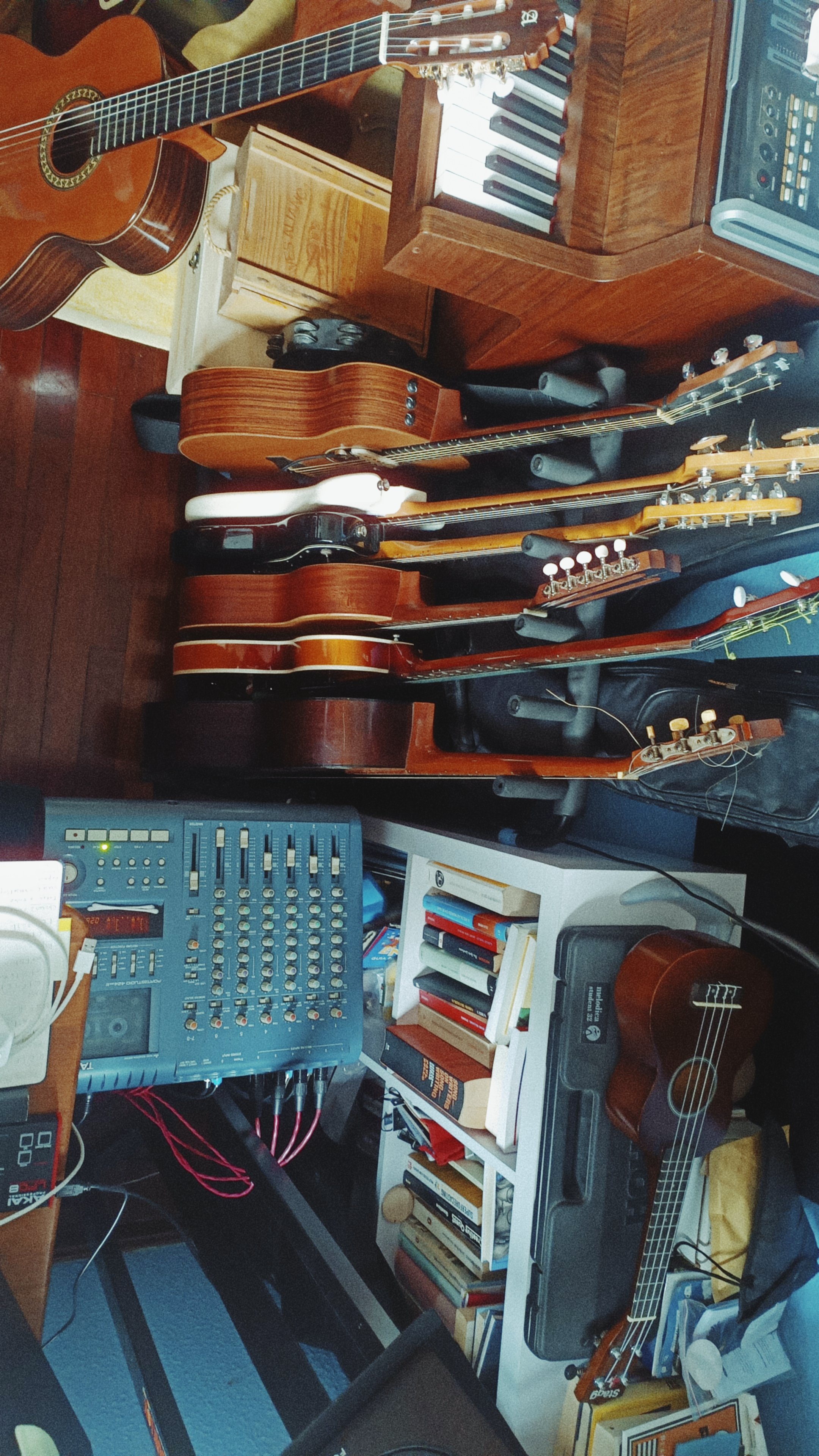 A room with musical instruments including several guitars, an electronic keyboard, and music equipment on a desk and shelves, along with books and electronic devices.