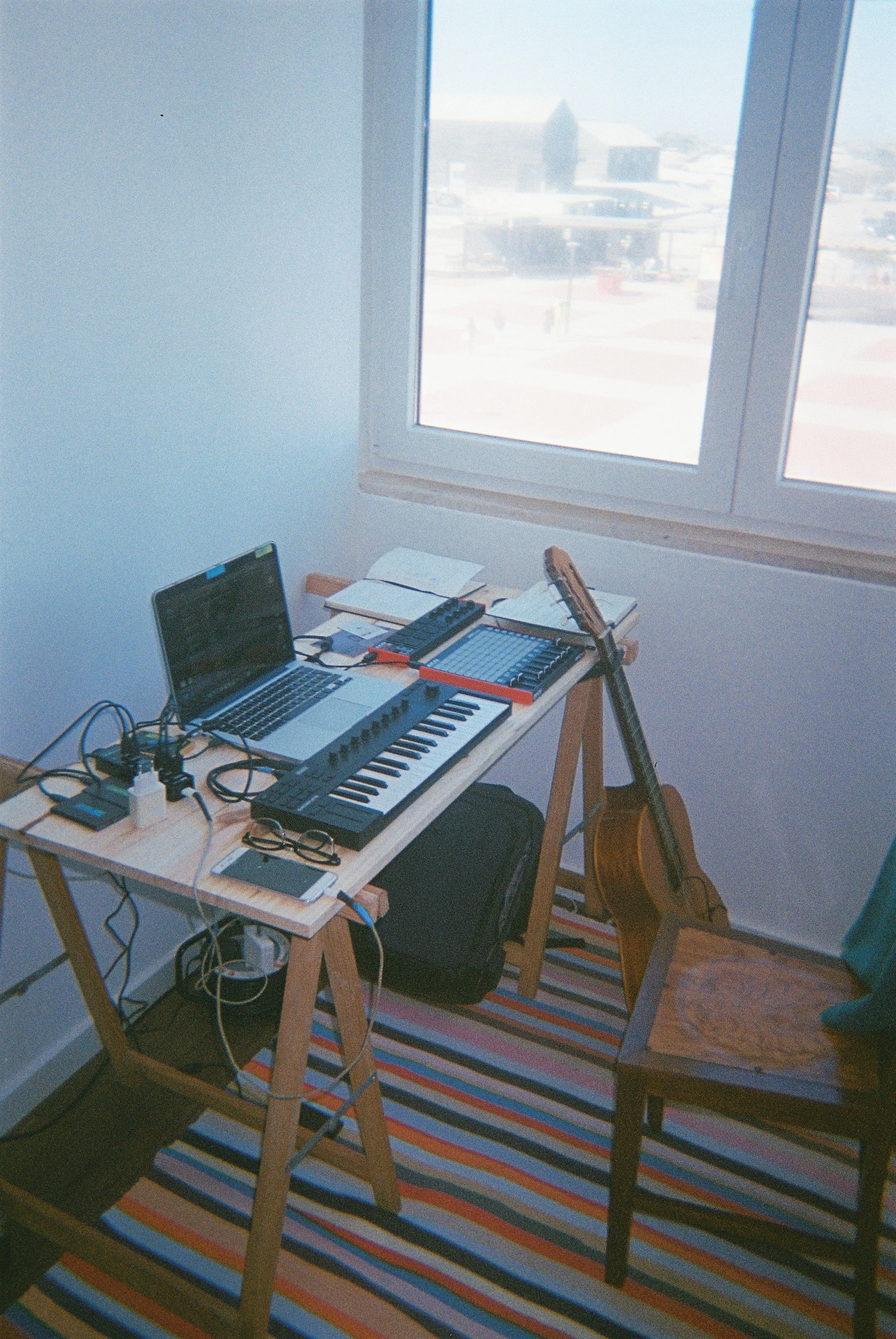 A music studio corner with a small wooden table holding electronic music equipment, a laptop, and a book, next to a guitar resting on a chair. The room has a window with a snowy landscape outside and a colorful striped rug on the wooden floor.