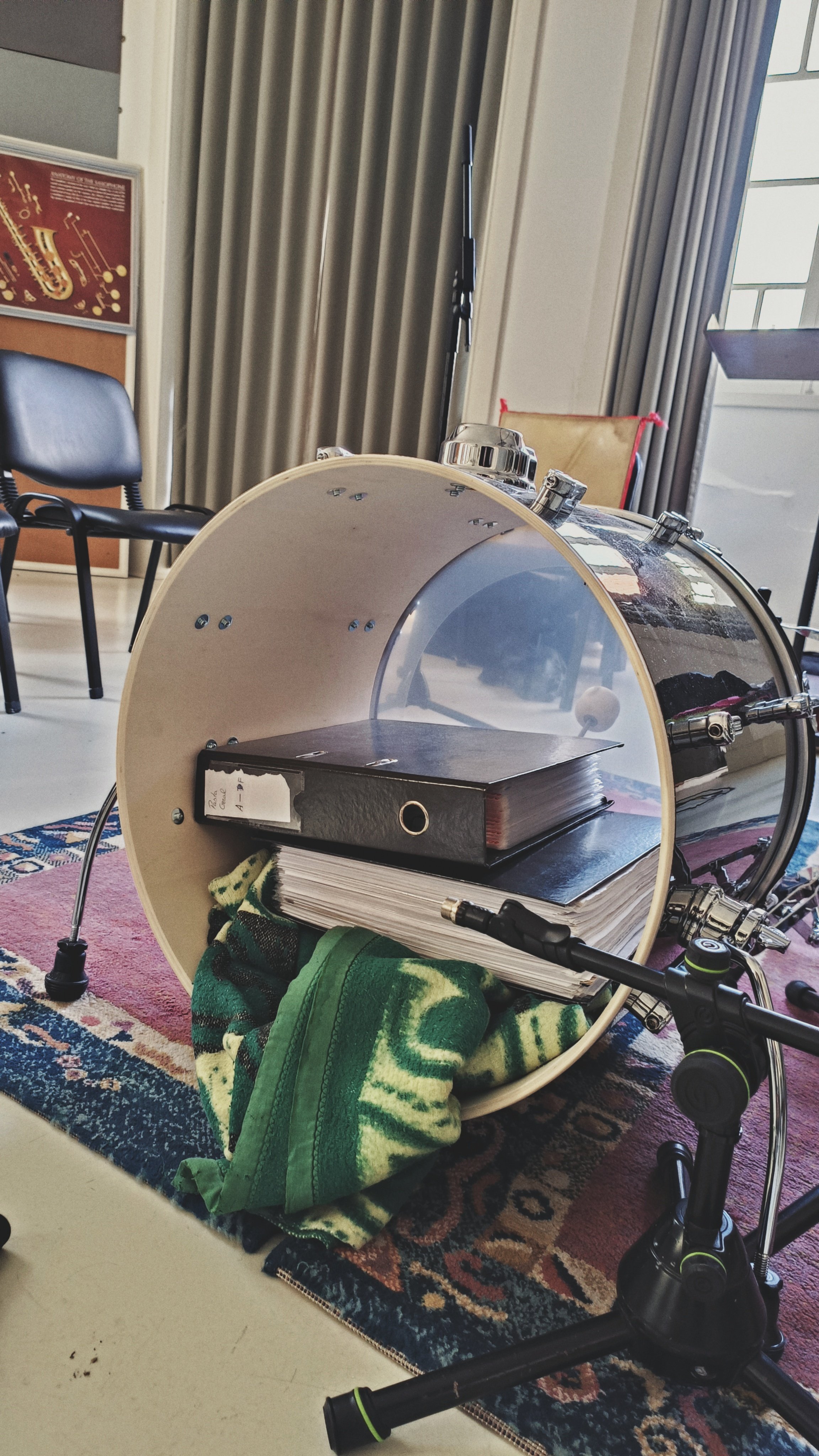 A drum set with a bass drum lying on its side, placed on a patterned rug with a green towel underneath, in a room with chairs, a window, and curtain.