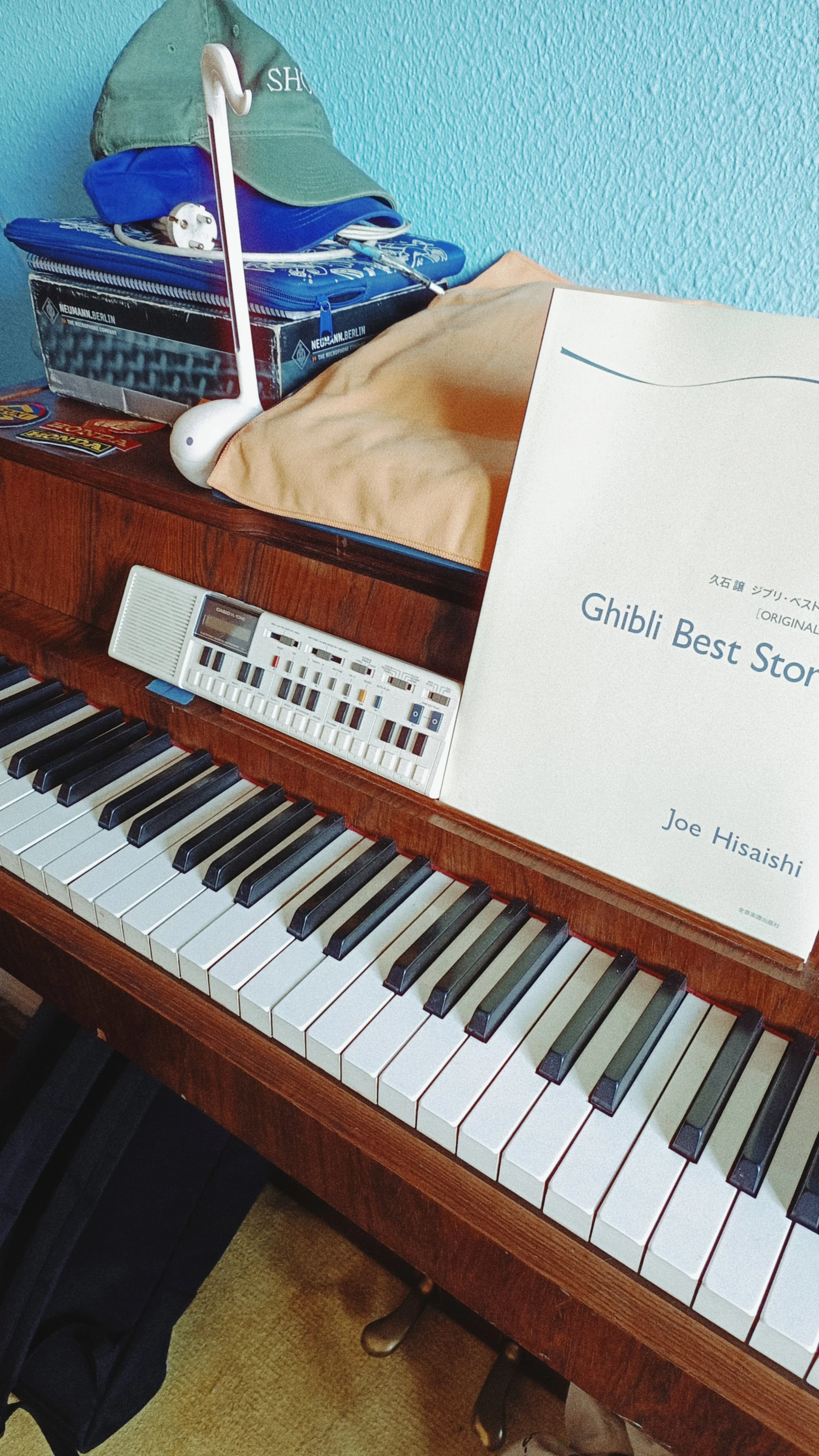 Close-up of a piano keyboard with a book titled 'Ghibli Best Songs' resting on the music stand. Items on top of the piano include a beige cloth, a box, a blue cap, and some electronic accessories.