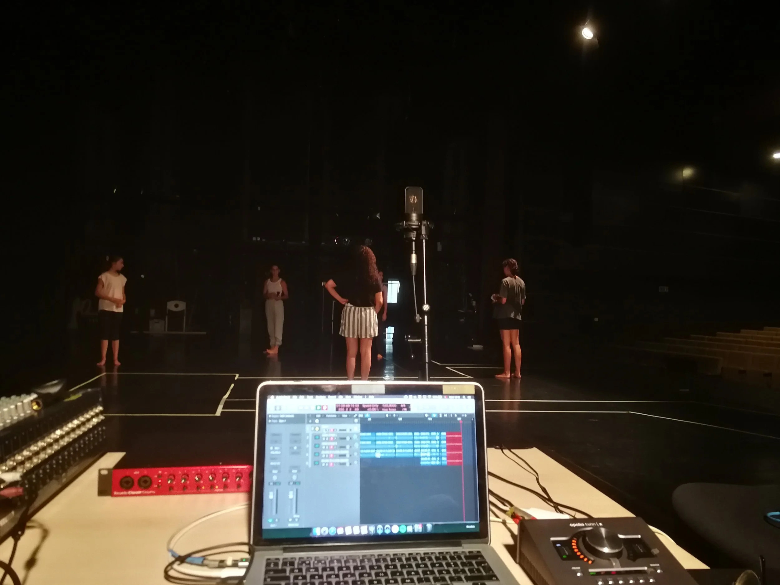 View from a music performance setup showing a laptop with DJ or music software, sound equipment, and a stage with four people standing and talking, with the night sky and bright moon above.