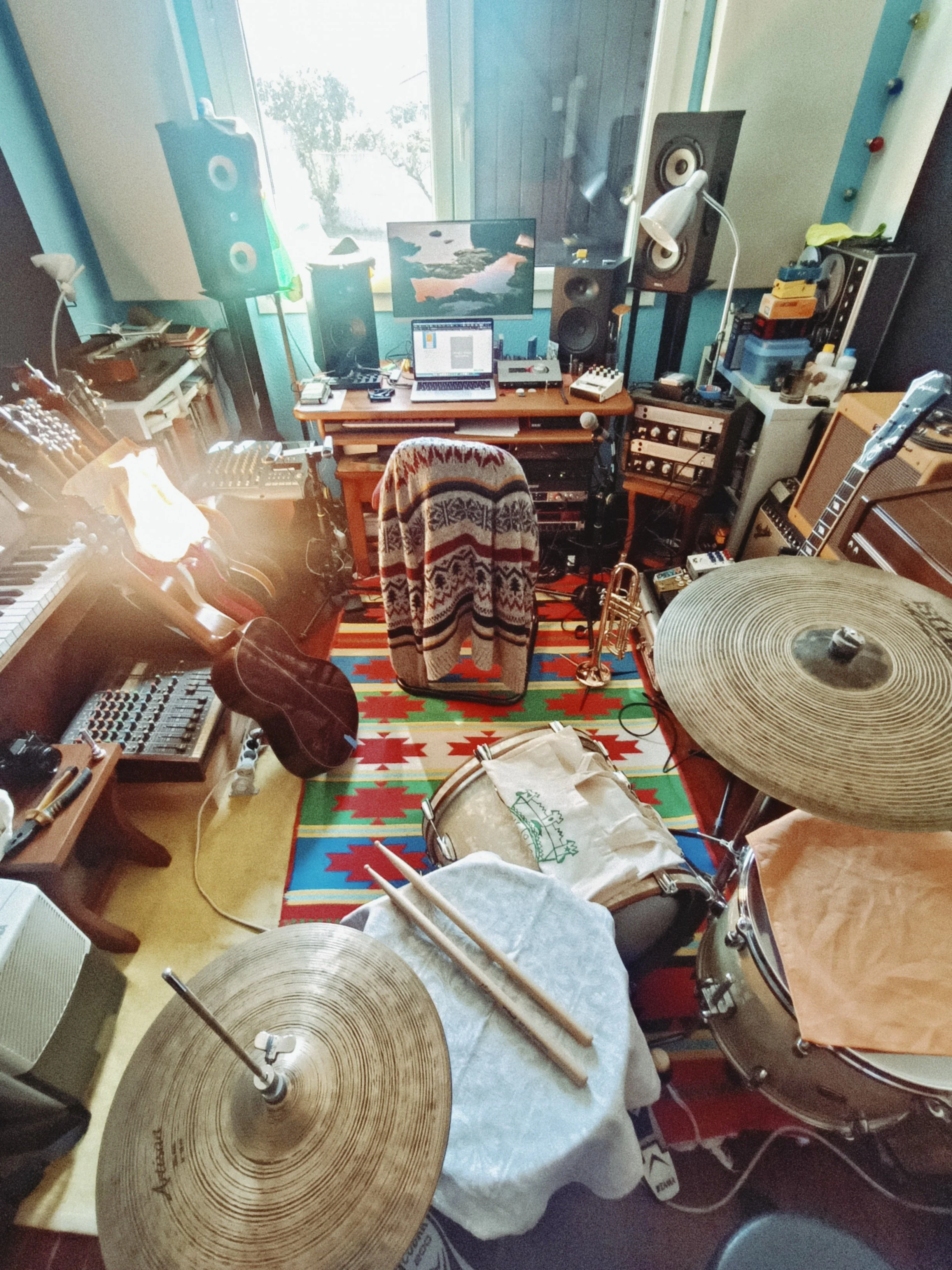A music studio with a drum set, guitars, keyboard, computer, and studio equipment. There are large cymbals in the foreground and various musical instruments and gear throughout the room.