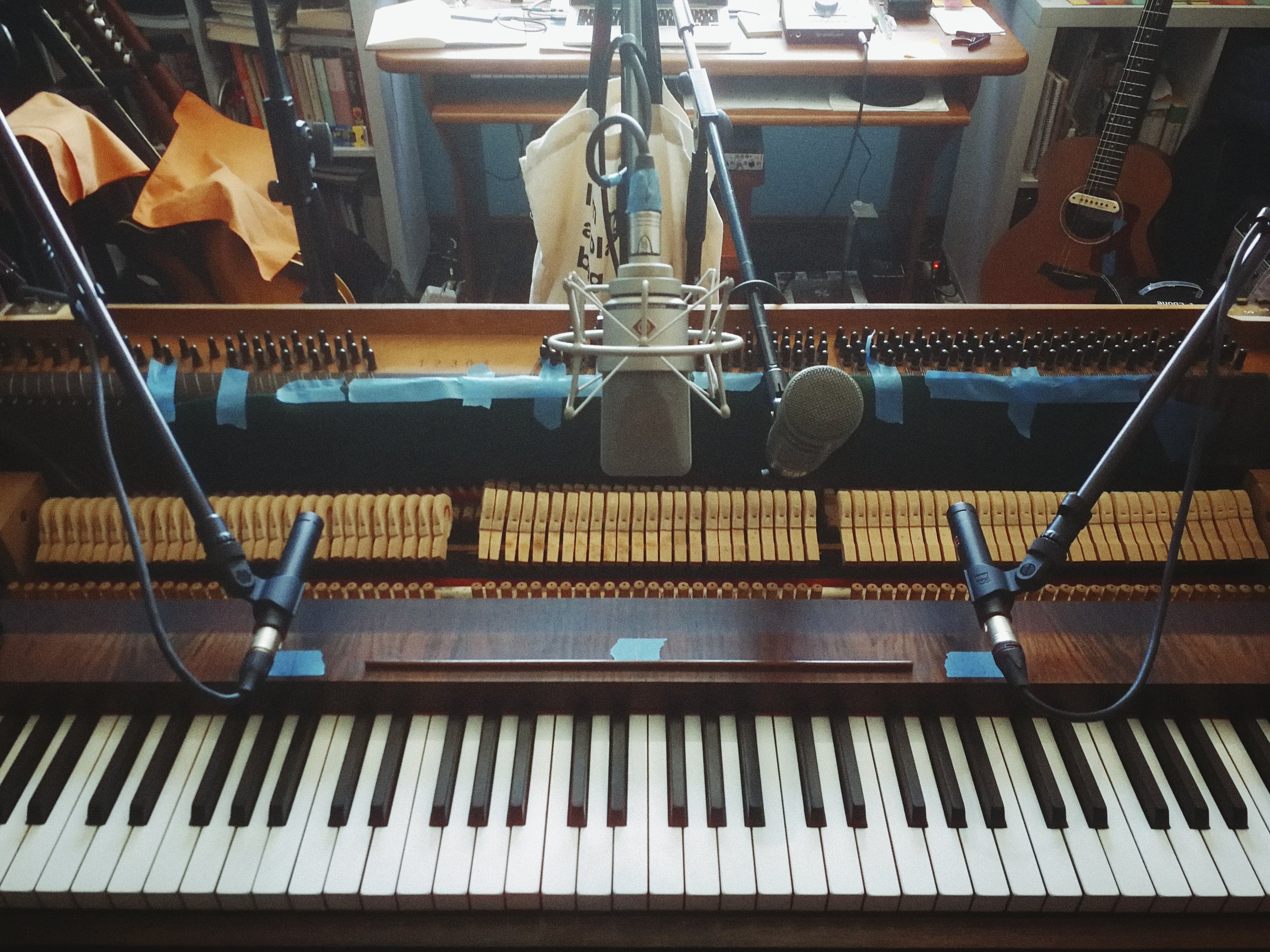 Close-up of an open piano with the inner strings and hammers visible, placed in a room with a bookshelf, guitars, and a desk in the background.