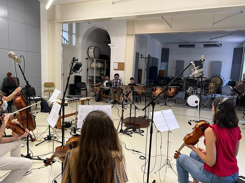 Orchestra rehearsal with musicians playing violins, cellos, and various percussion instruments in a recording studio.
