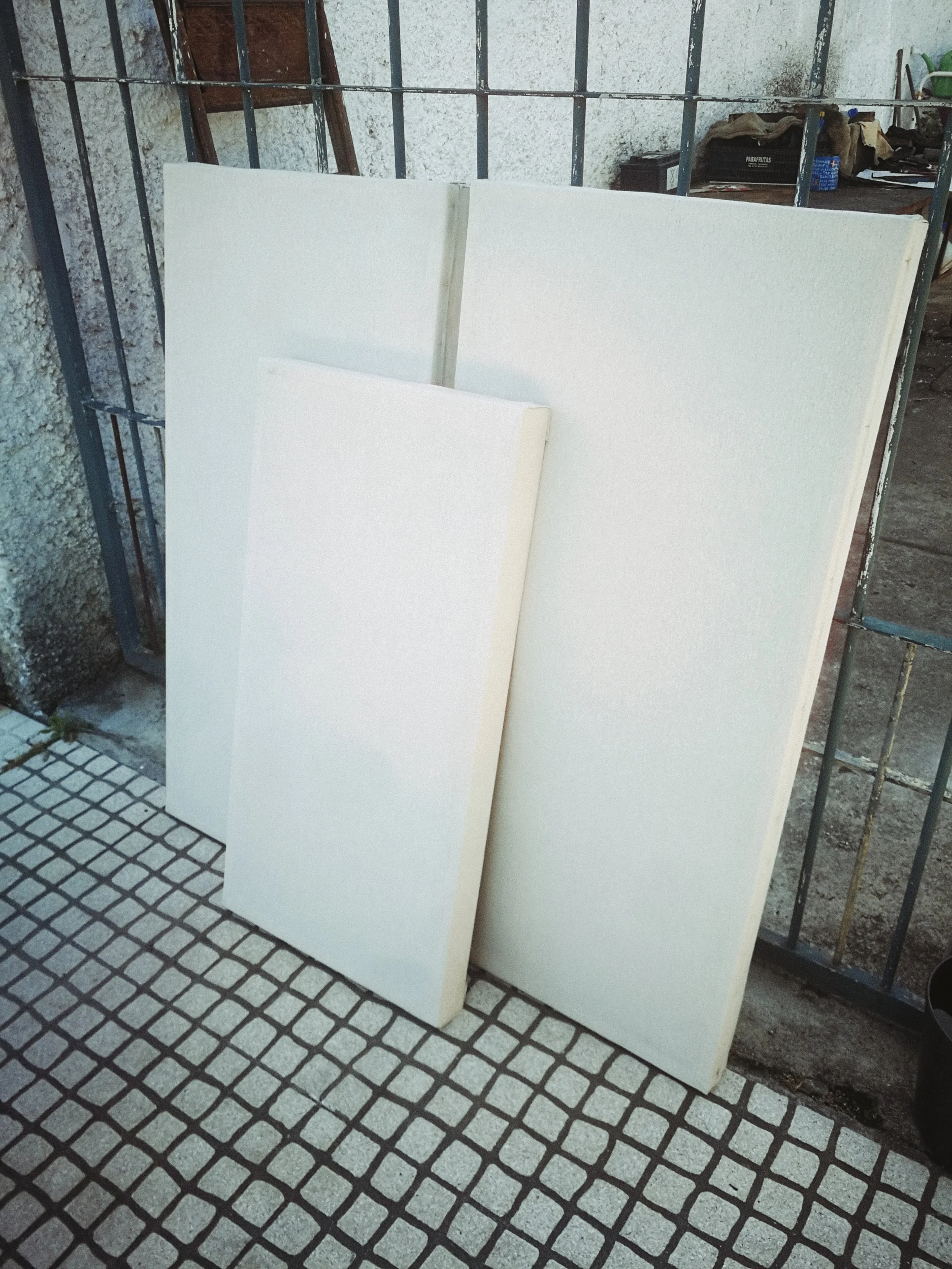 Two large white foam boards leaning against a metal fence on a tiled sidewalk with a textured wall and some miscellaneous objects in the background.