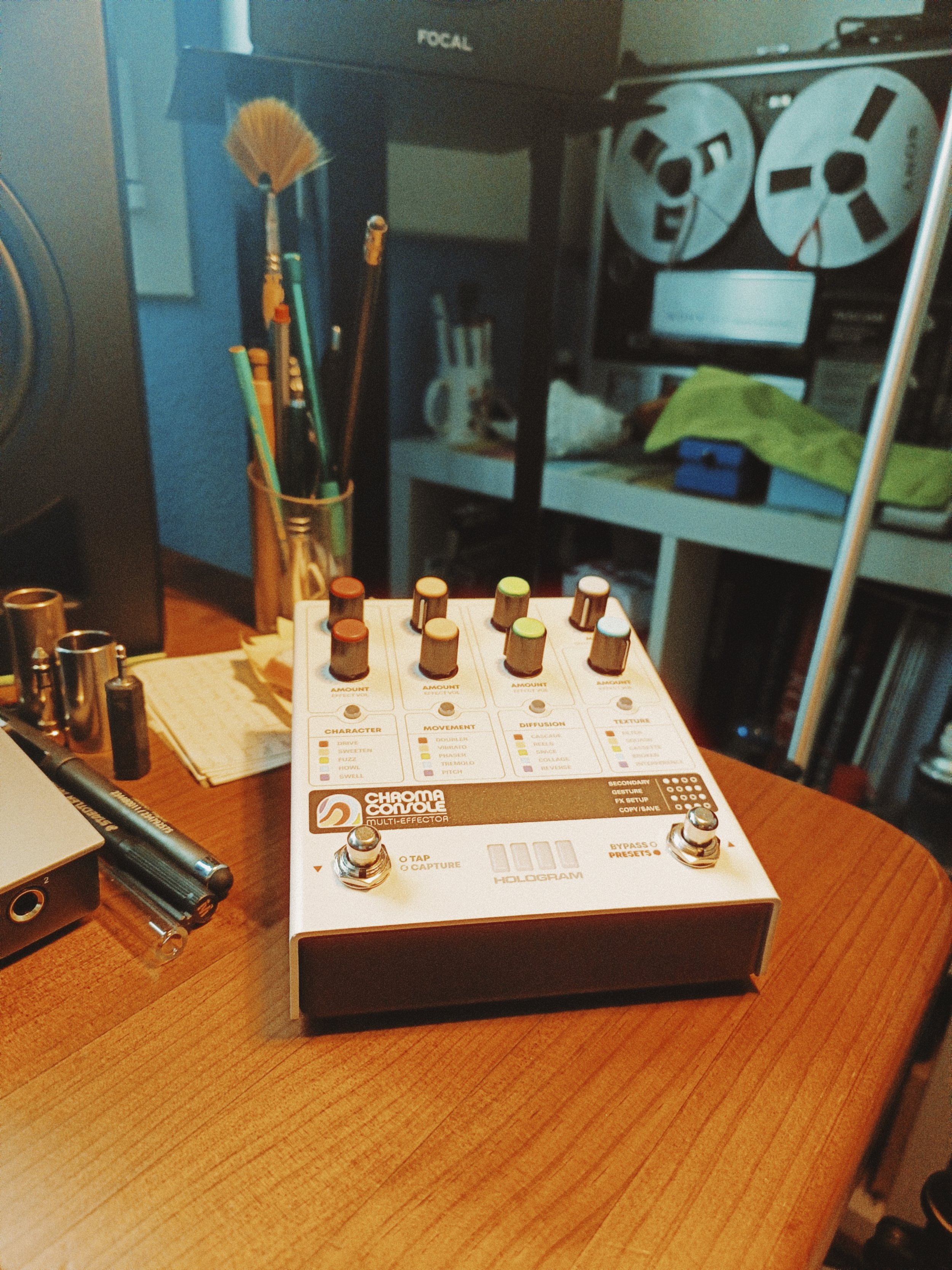 A musical effects pedal labeled 'Chama Console' with multiple knobs, foot switches, and a small display, placed on a wooden table. Surrounding objects include a black marker, small cylindrical objects, and a background with a desk, books, and electro