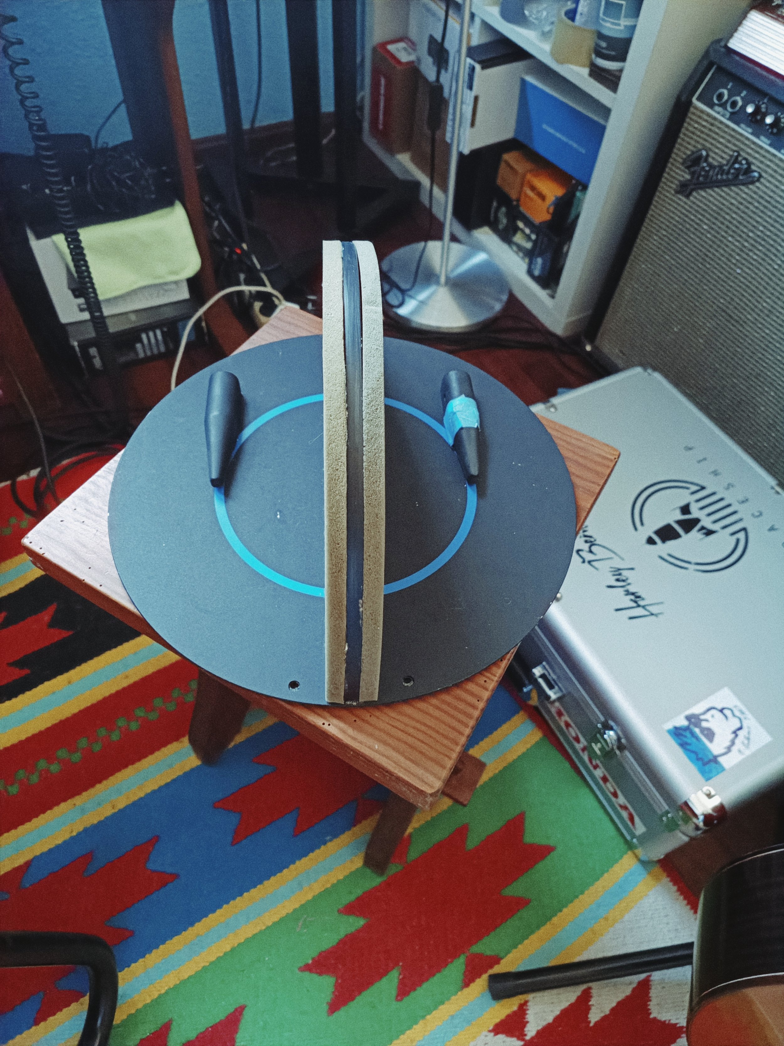 A ping pong paddle on a round black table with a blue circle, placed on a small wooden table on a colorful, striped red, green, blue, and yellow rug.