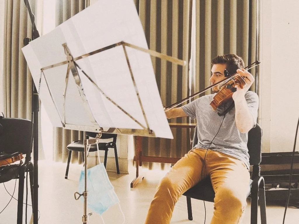 A man playing a violin in a music studio, sitting on a chair, with sheet music on a stand in front of him.
