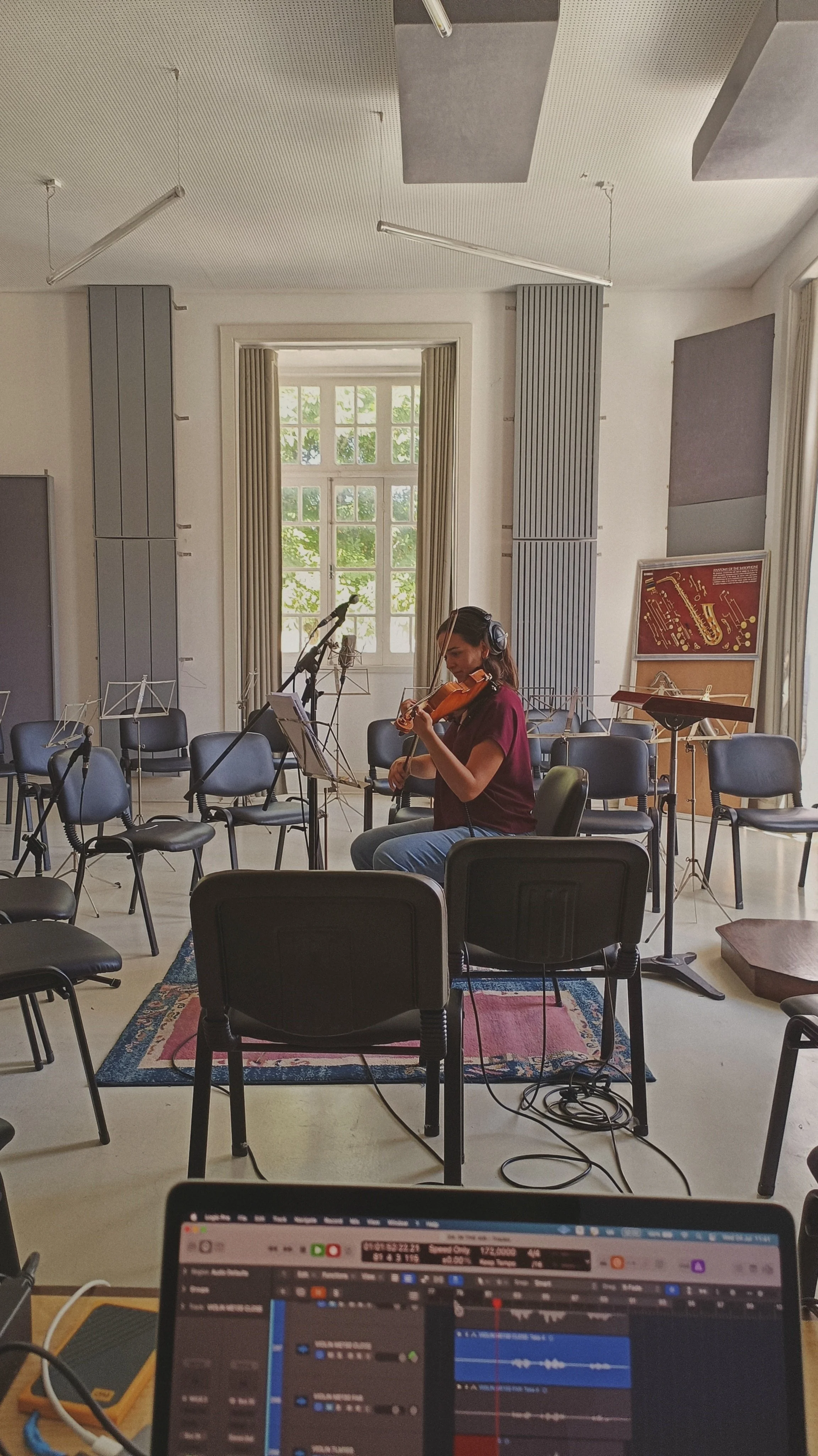A woman playing the violin in a music studio with large windows and music stands, while a computer monitor and recording equipment are visible in the foreground.