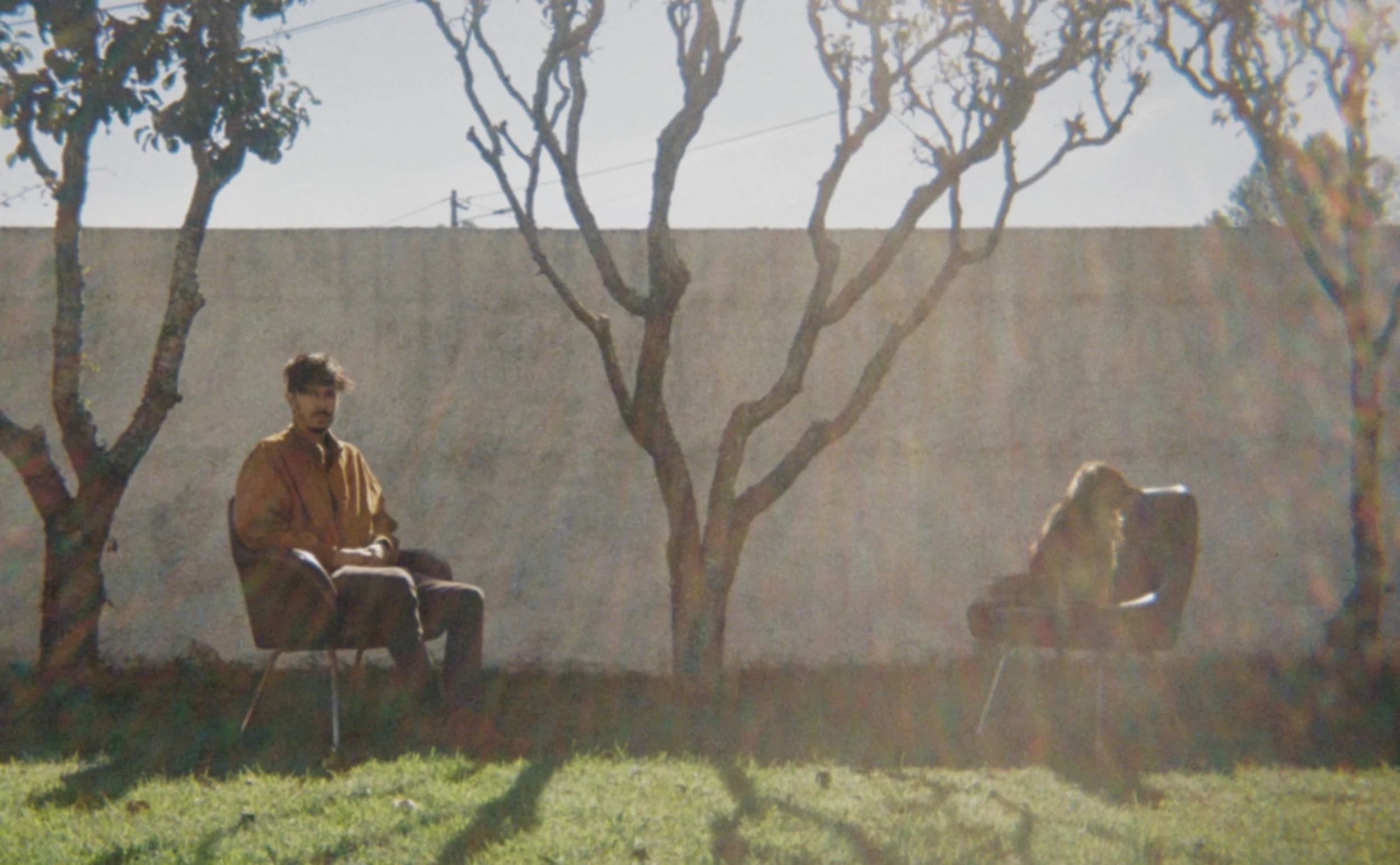 A man with dark hair, beard, and glasses sitting on a chair outdoors under leafless trees with sunlight shining through, in front of a tall concrete wall.
