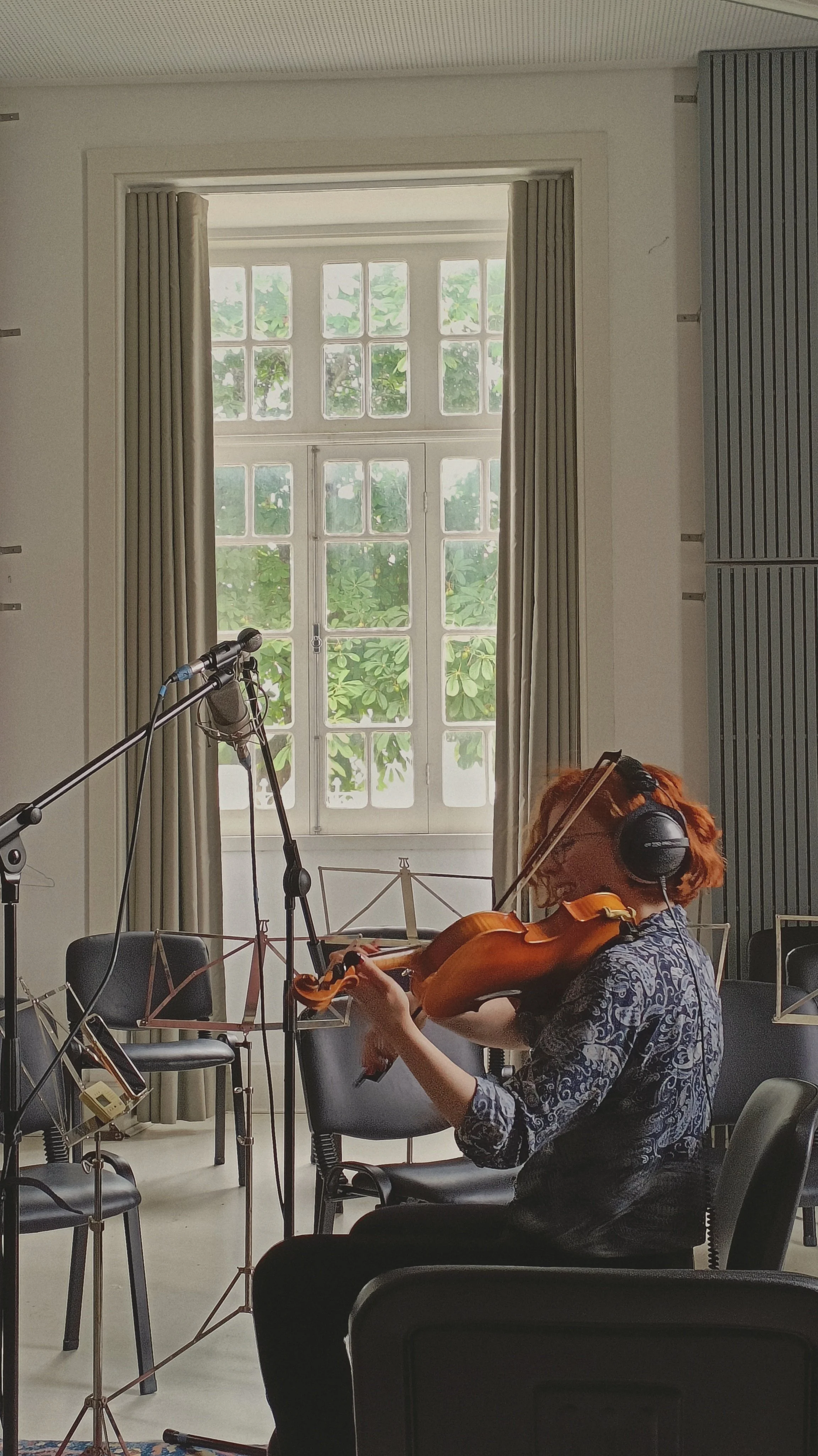 A woman is playing the violin in a room with a large window. She is wearing headphones and a patterned shirt, sitting on a chair, and there are music stands and chairs around her.