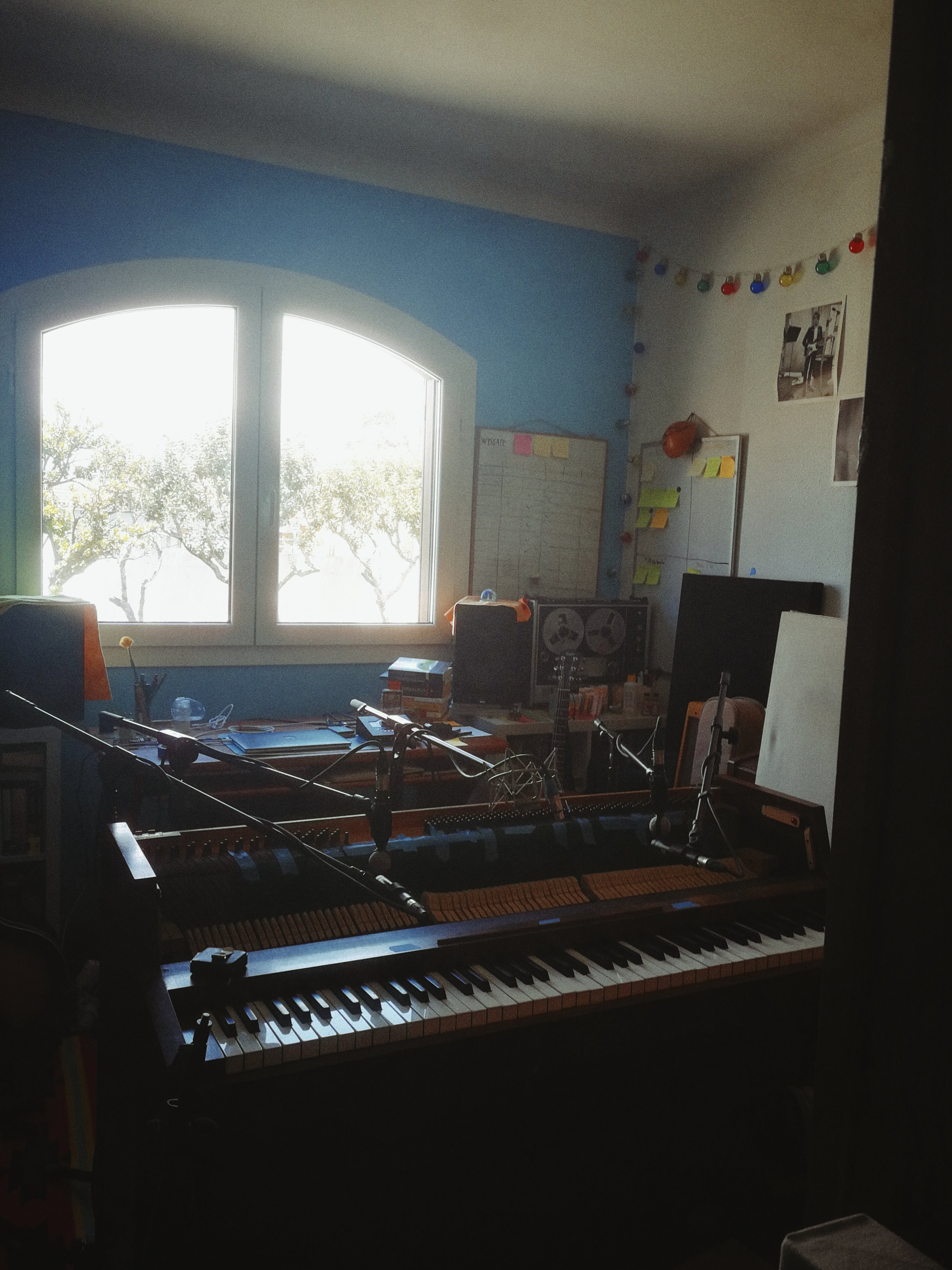 Indoor room with a grand piano, computer monitor, and cluttered desk near a window with trees outside. The walls have colorful string lights and photos.