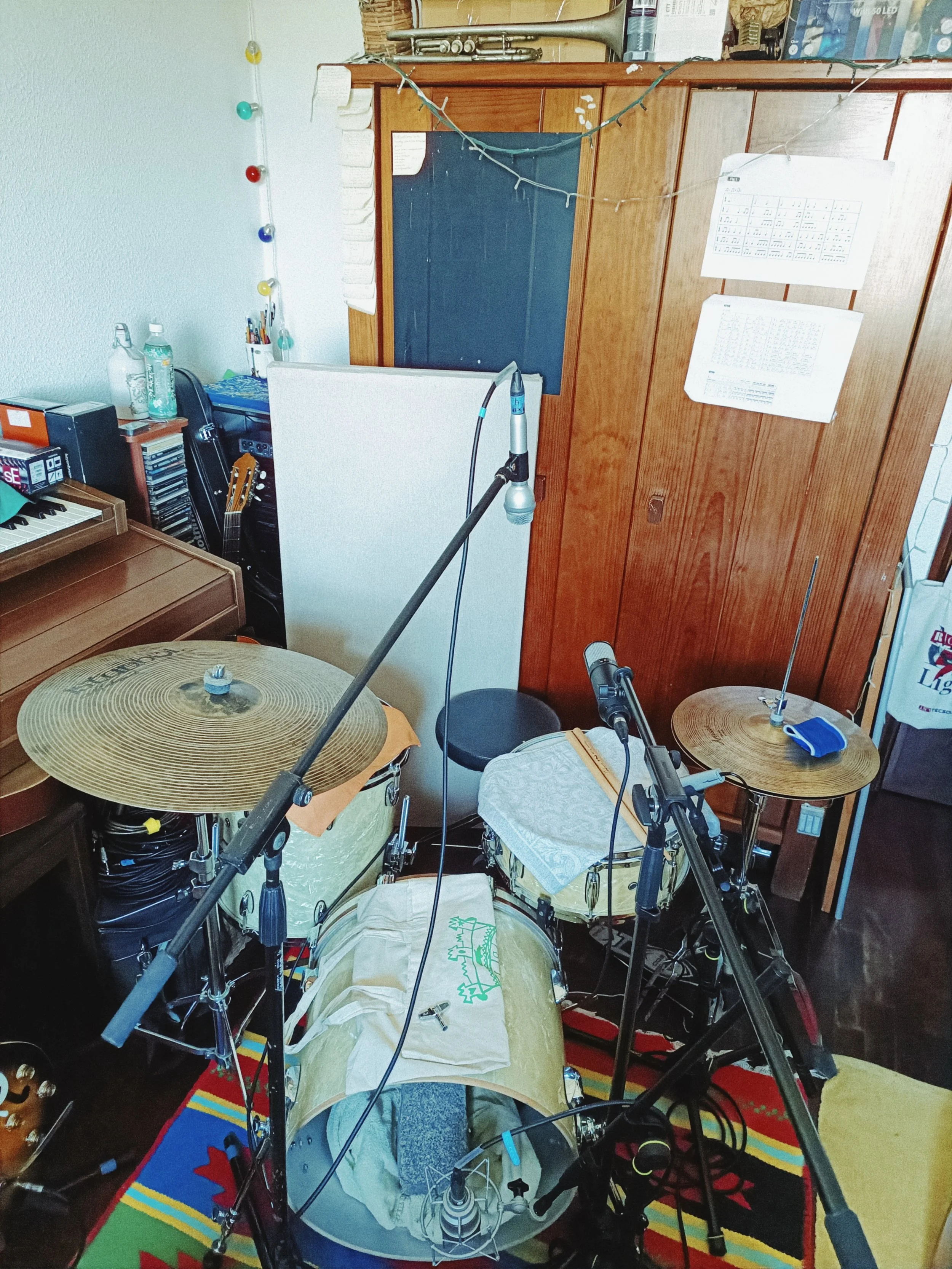 A room with musical instruments, including a drum set and cymbals, with sheet music and microphone stands, and a colorful rug on the floor.