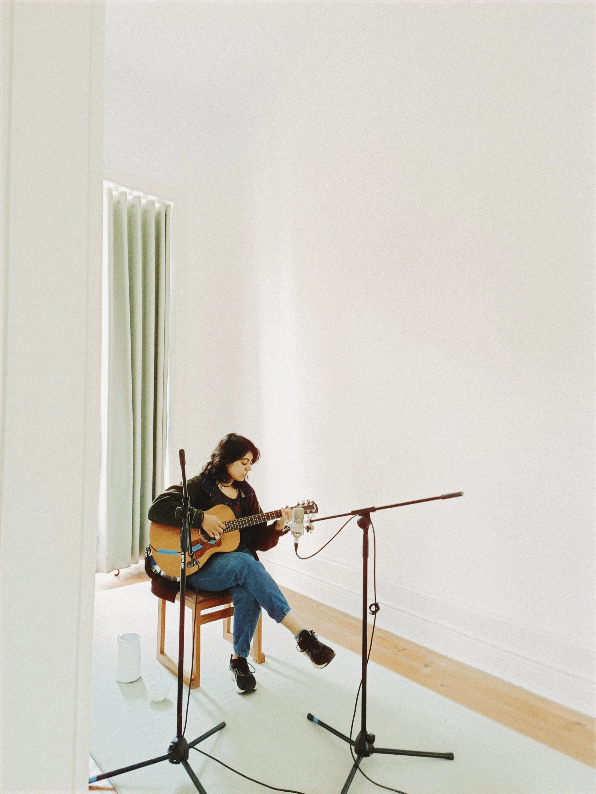 A woman sitting on a wooden chair in a bright, minimalist room playing an acoustic guitar while being recorded with multiple microphones, with large windows and white curtains.