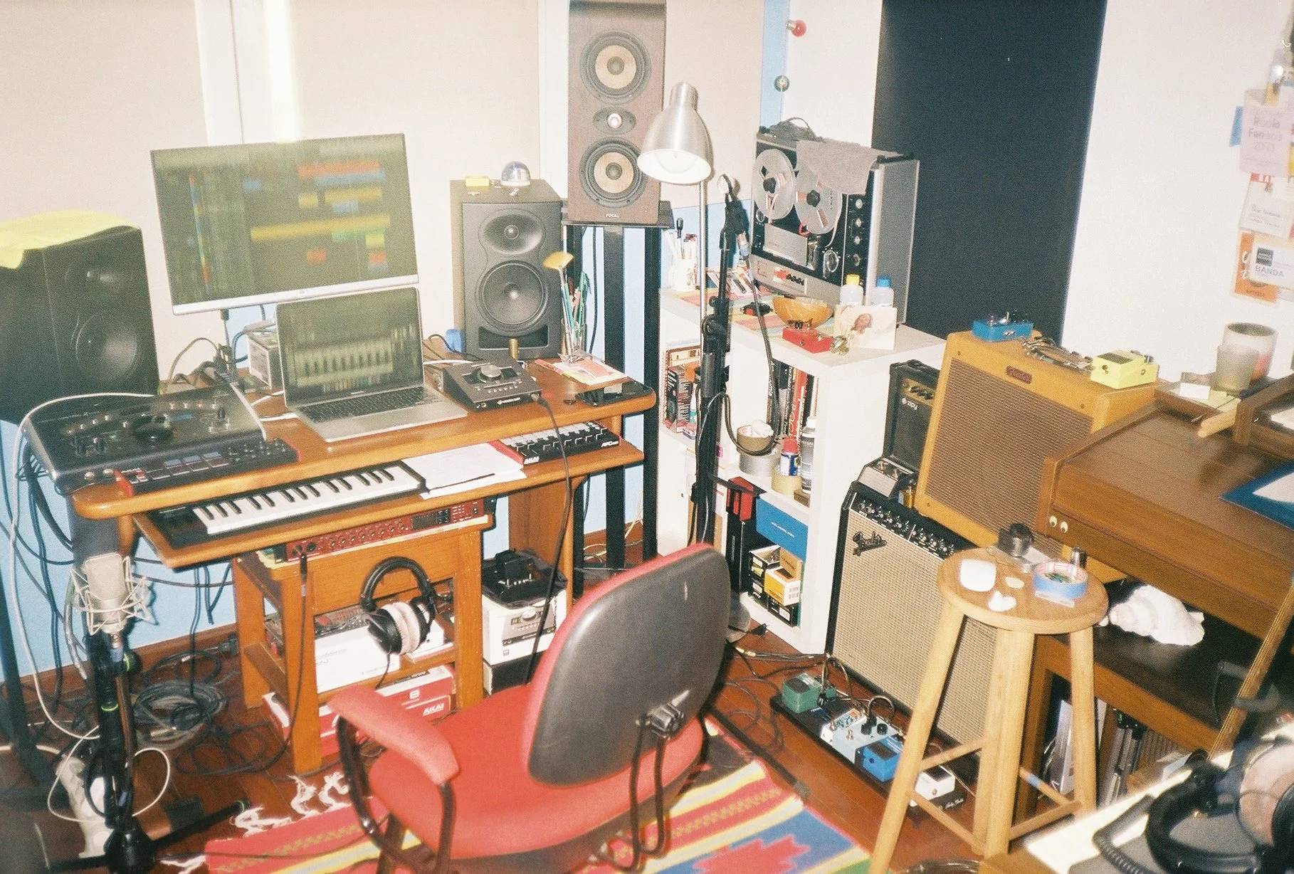 A music studio with various instruments and equipment including a computer, keyboard, speakers, guitar amps, and a microphone. There's a small wooden table with candles and a stool beside it.