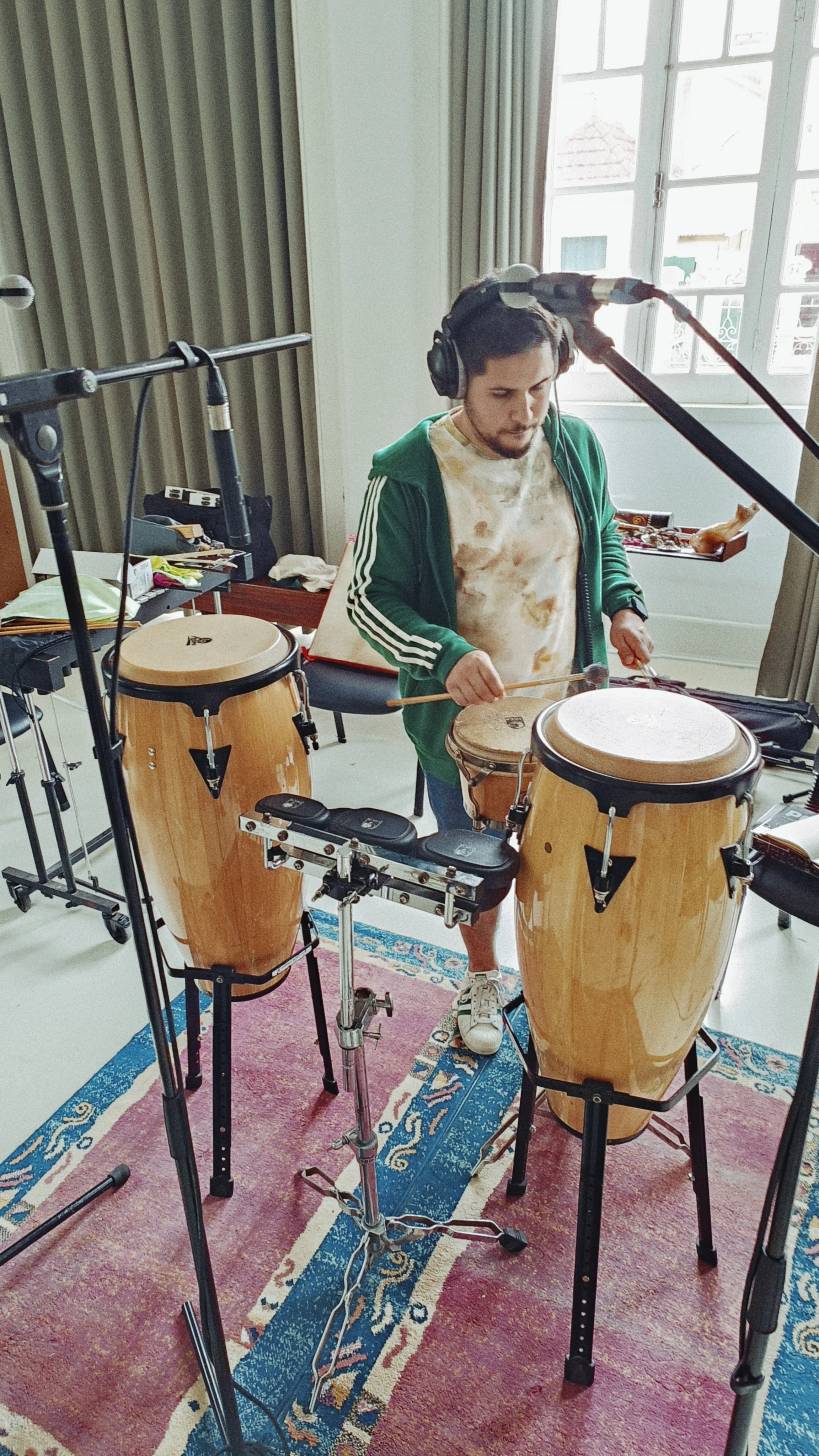A man playing conga drums and a percussion setup in a room with large windows, surrounded by microphone stands and musical equipment.
