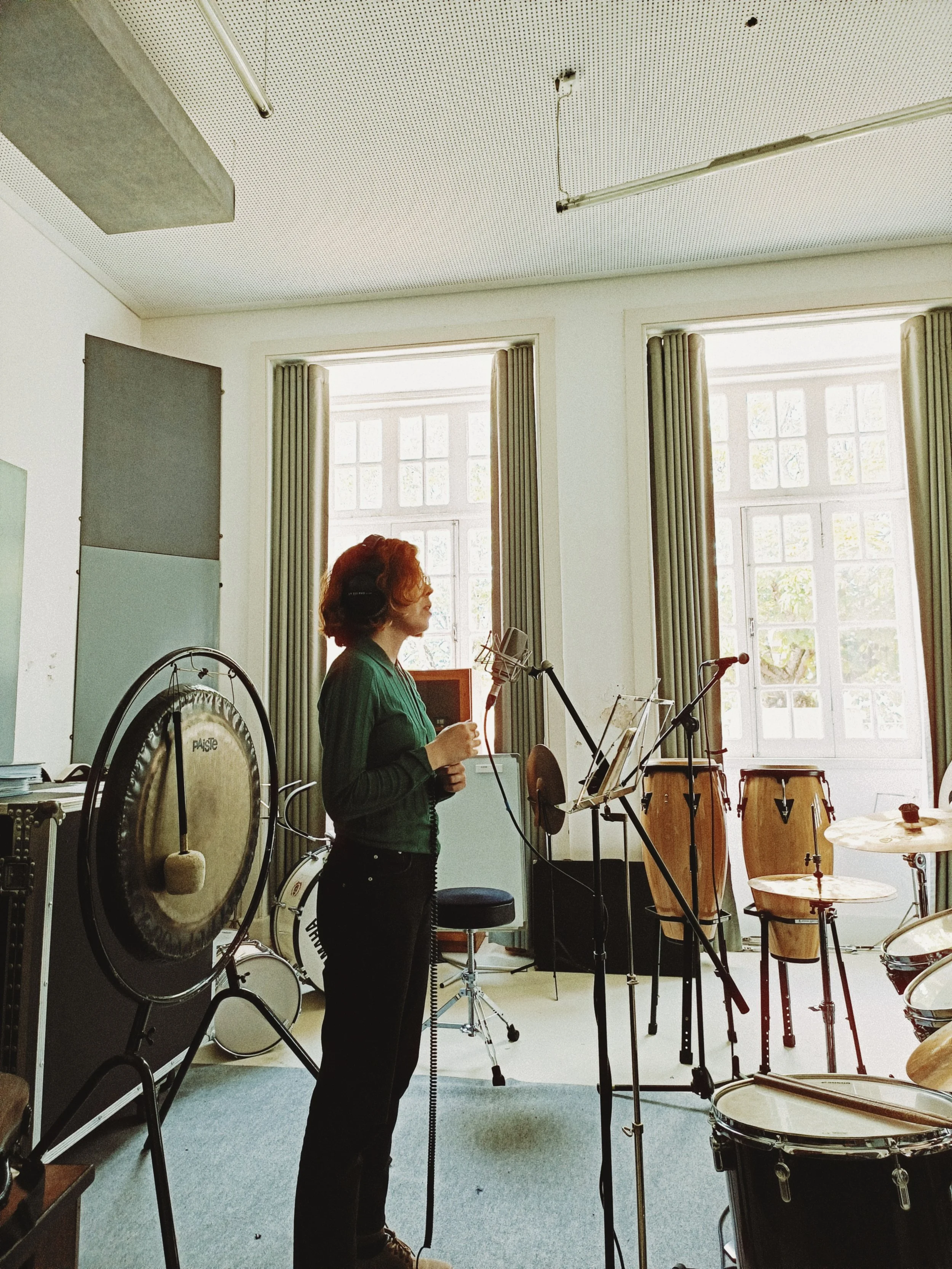 A woman with red hair and glasses standing in a music studio, wearing headphones and holding a microphone, surrounded by various percussion instruments including a gong and drums, with large windows and music equipment in the background.
